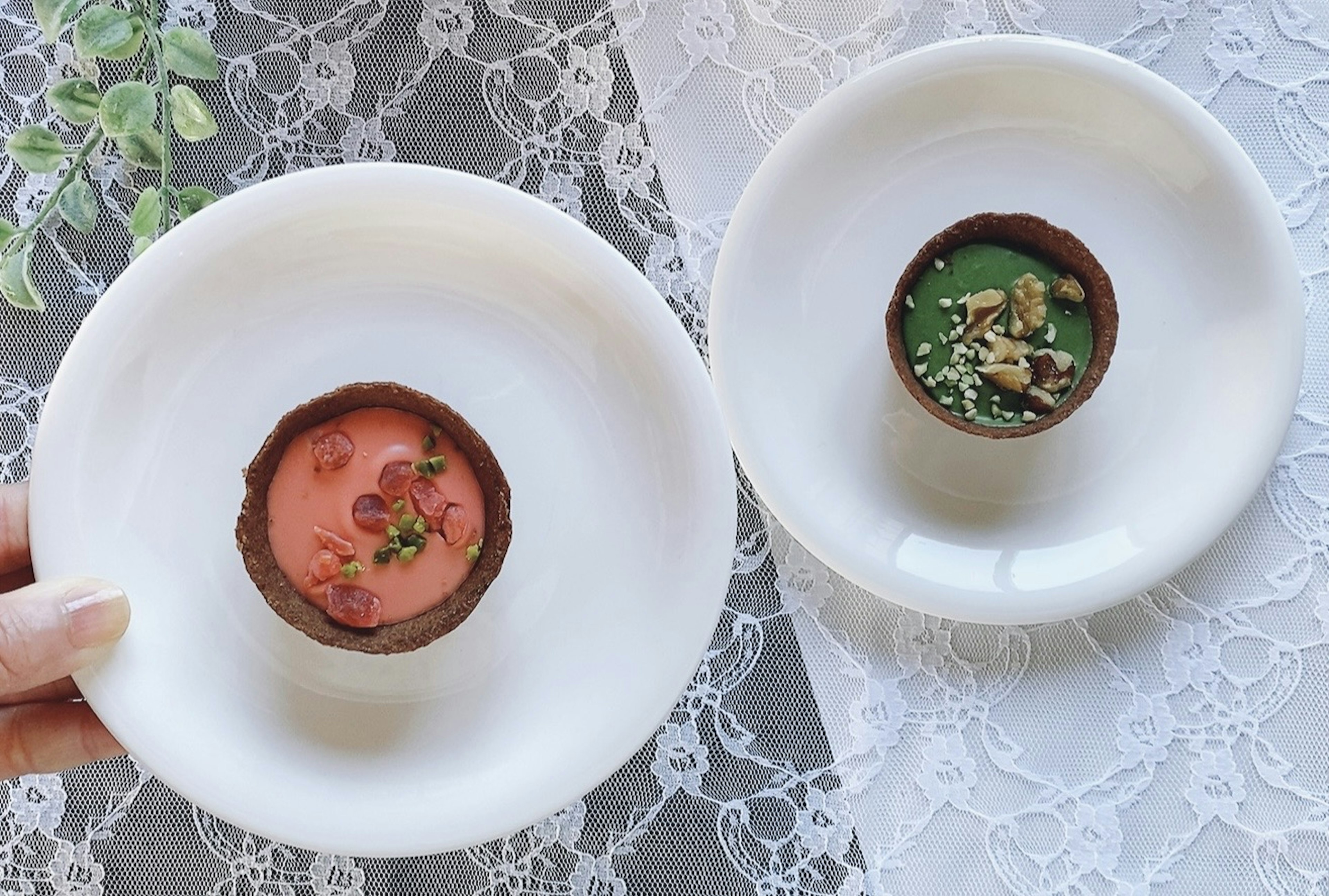 Dos postres en platos blancos sostenidos con una mano Uno es rosa con coberturas El otro es verde con nueces y hierbas