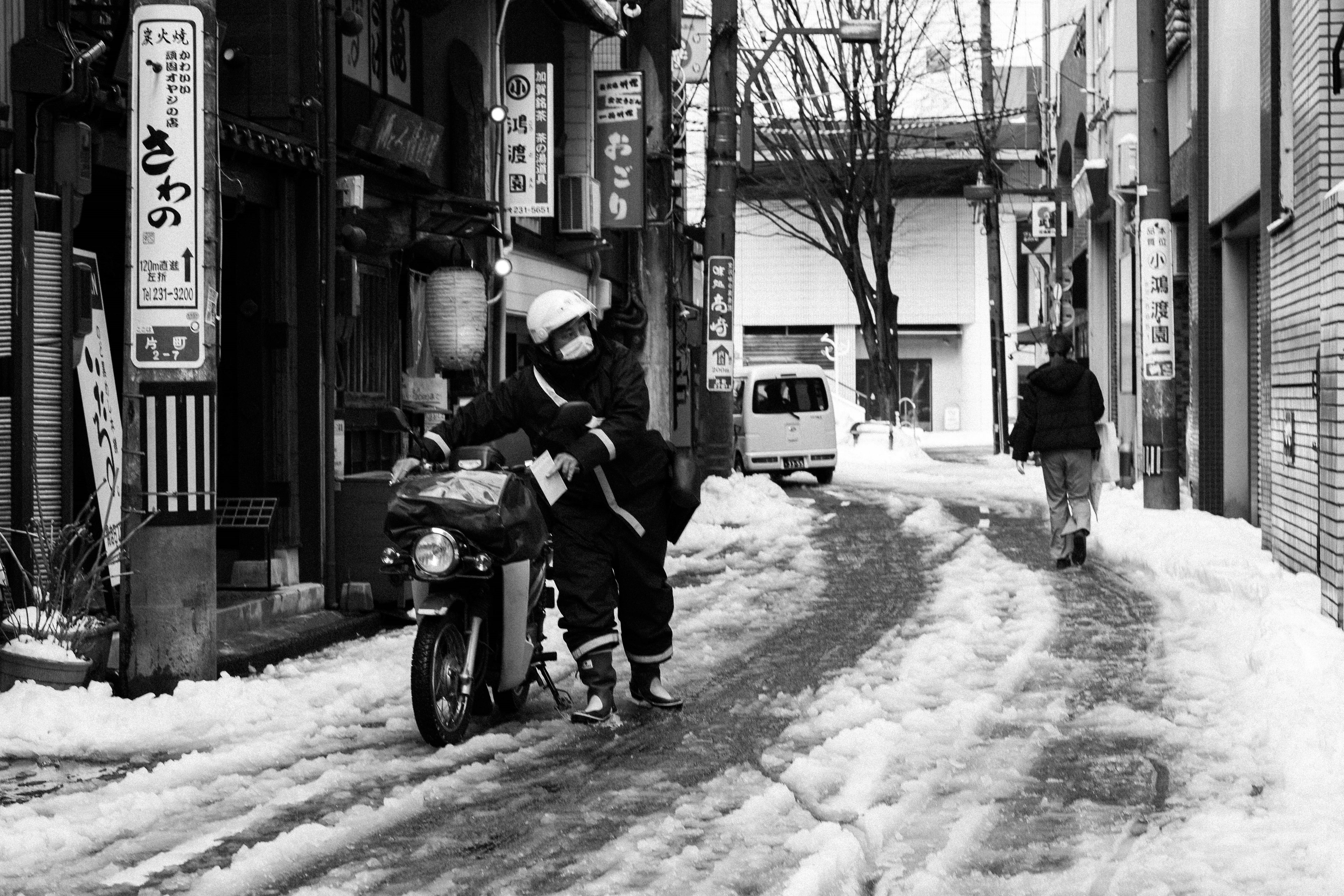 雪に覆われた狭い路地を走るバイクと歩行者