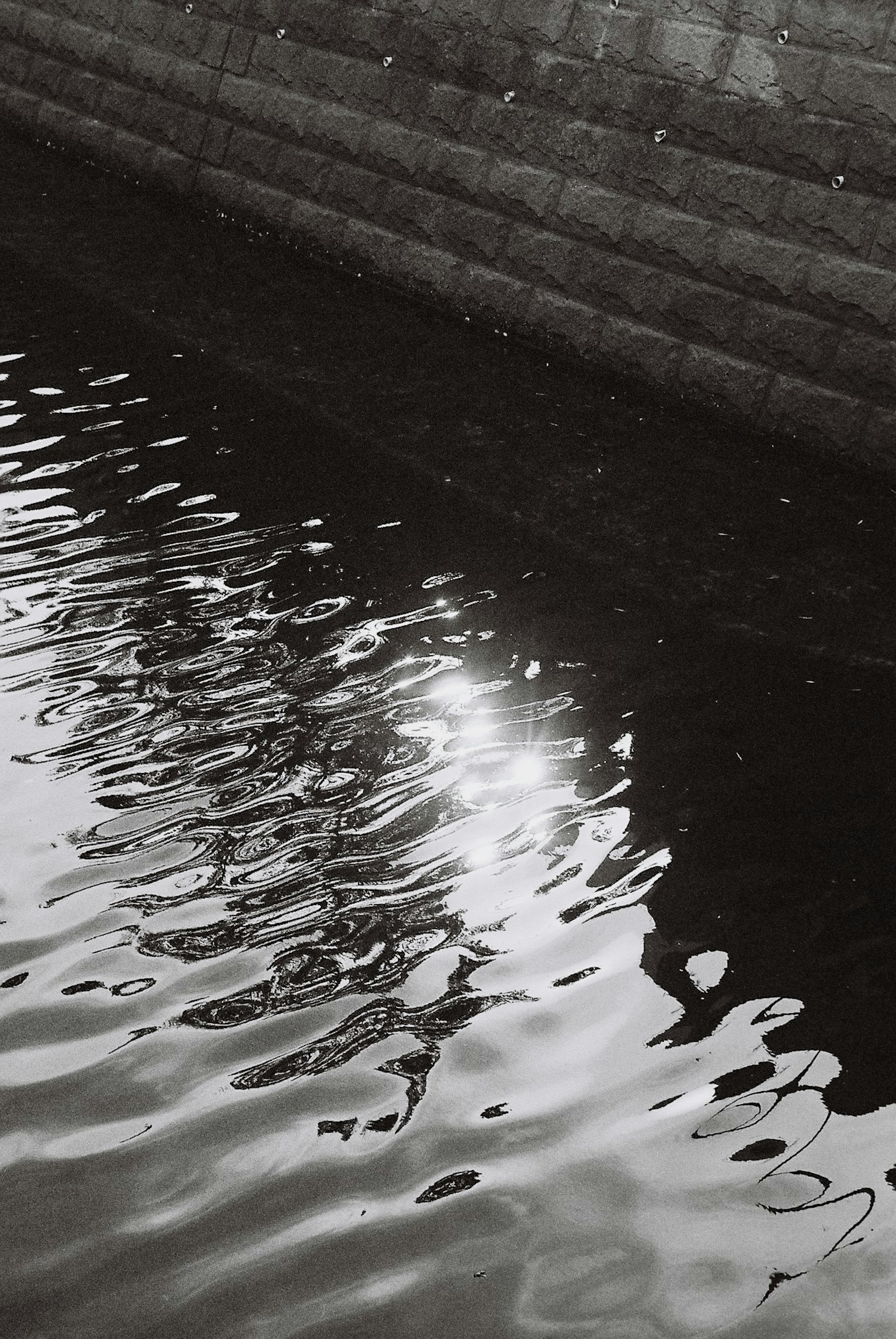 Surface d'eau en noir et blanc reflétant la lumière et un mur en pierre