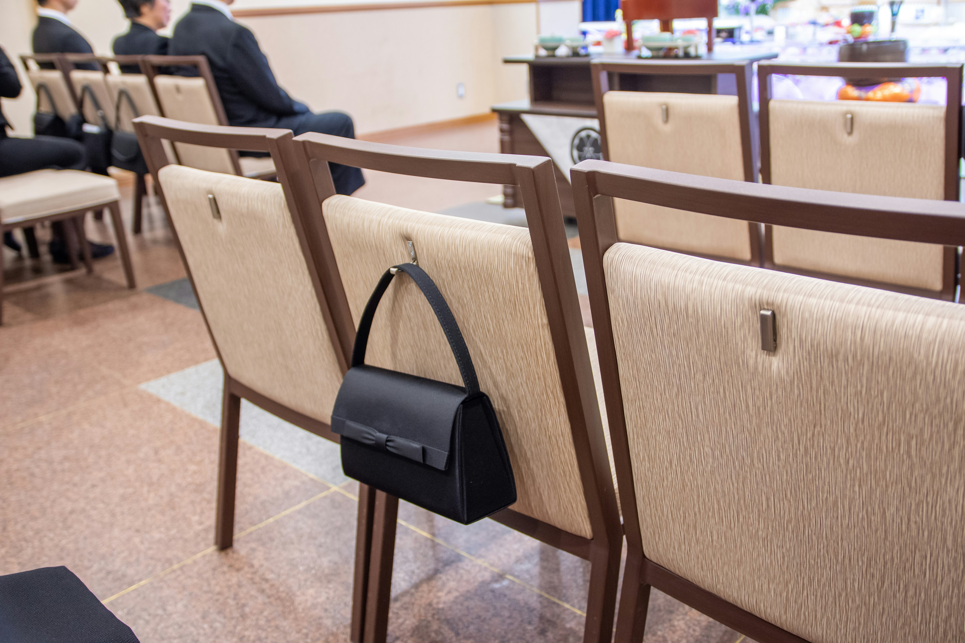 Eine schwarze Handtasche, die an der Rückenlehne eines Stuhls in einem formellen Umfeld hängt, mit weiteren Stühlen im Hintergrund