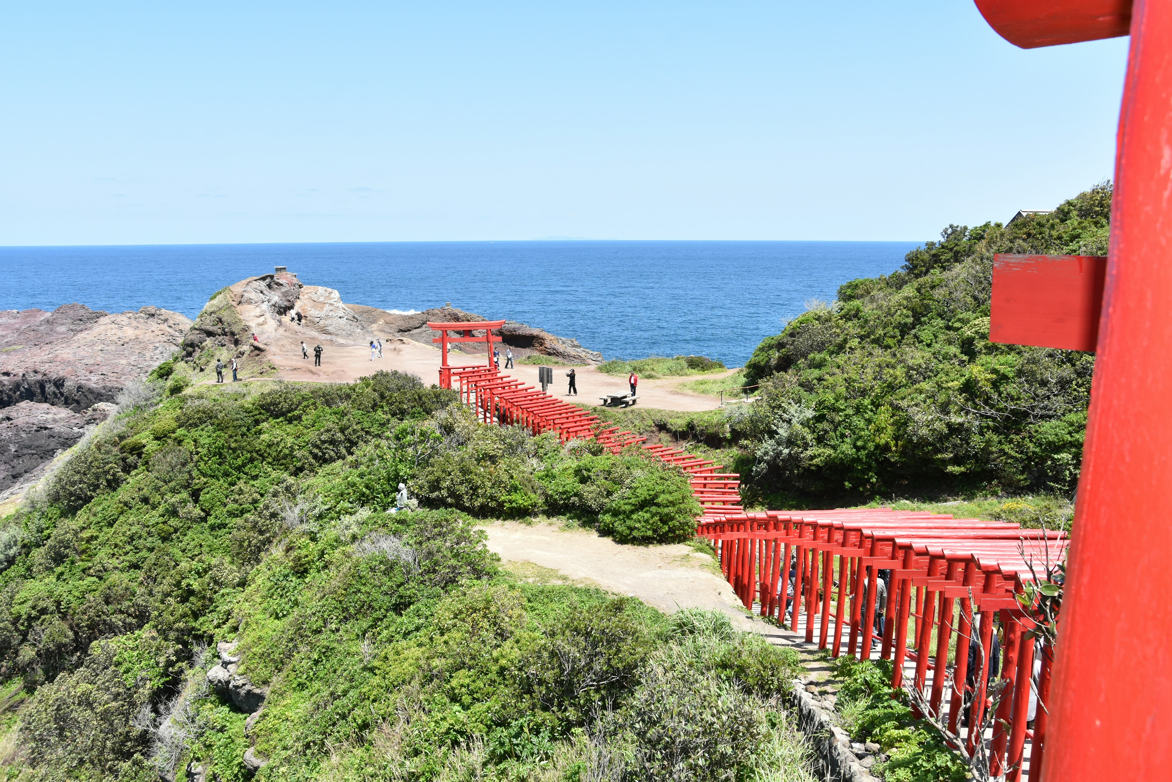 A path lined with red torii gates overlooking the blue ocean and green hills