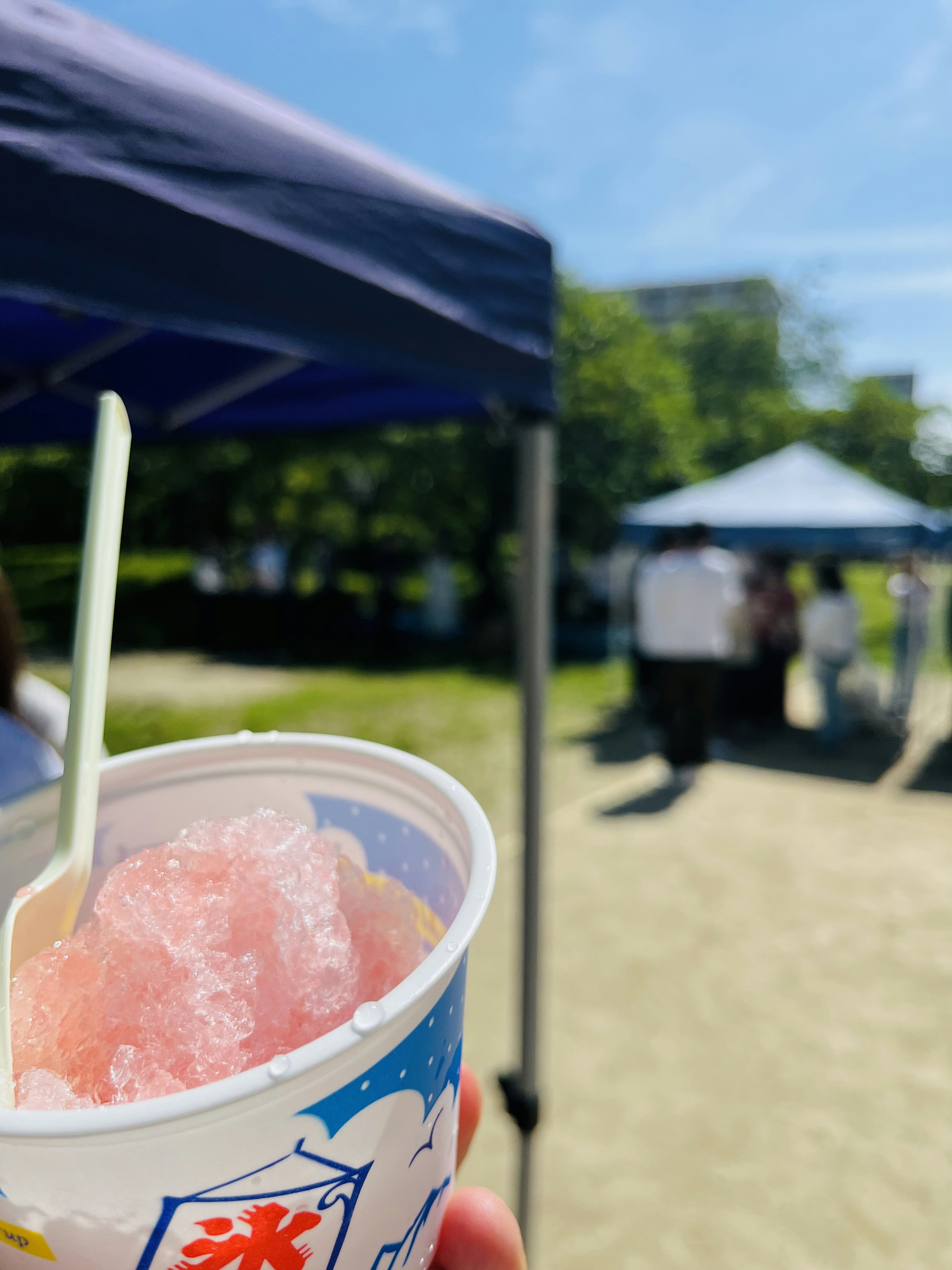 Eine Hand hält einen Becher mit Shaved Ice unter einem blauen Himmel mit Zelten im Hintergrund