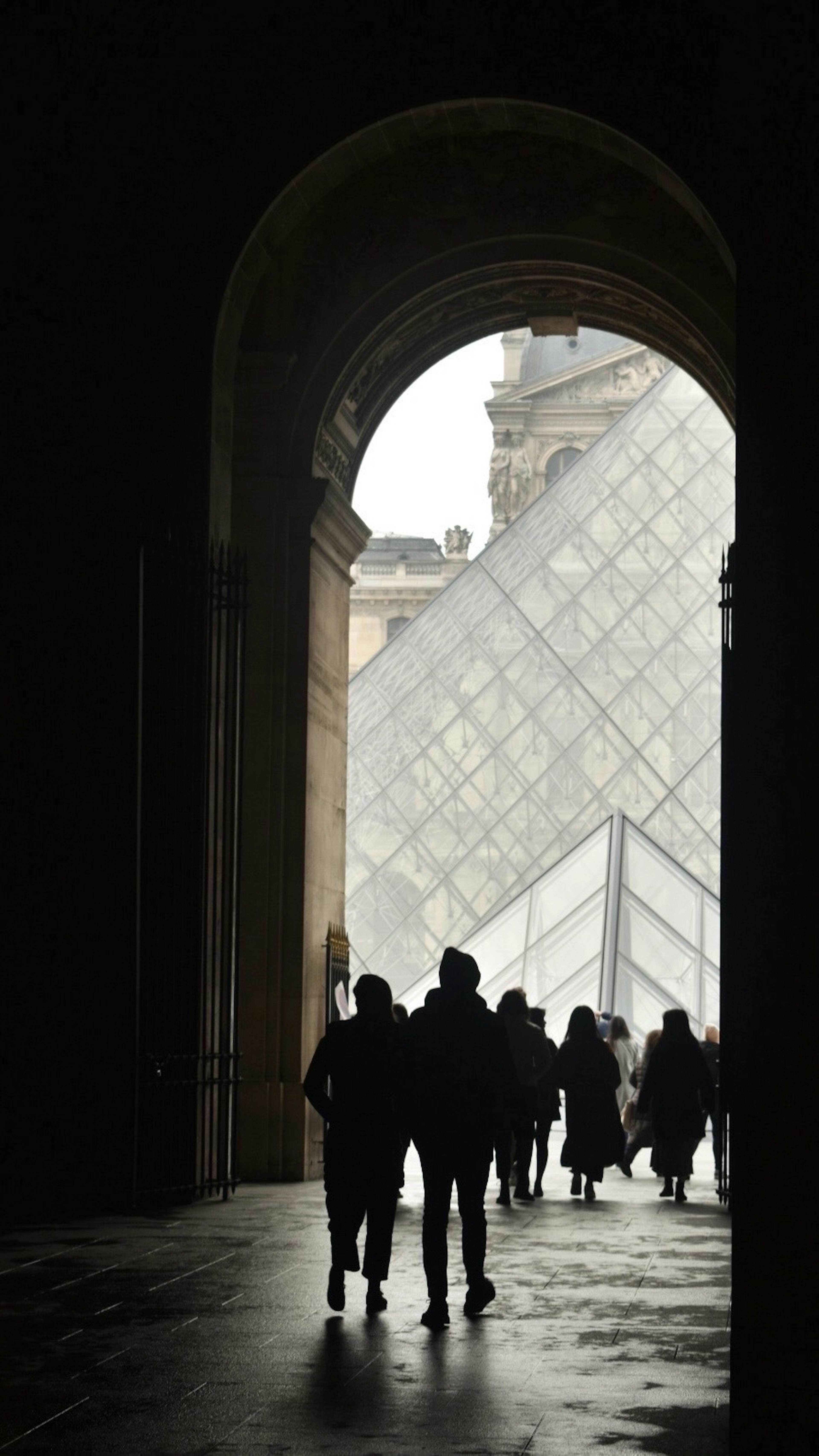 Silhouette của mọi người đi qua lối vào hình vòm của Bảo tàng Louvre với kim tự tháp kính nhìn thấy