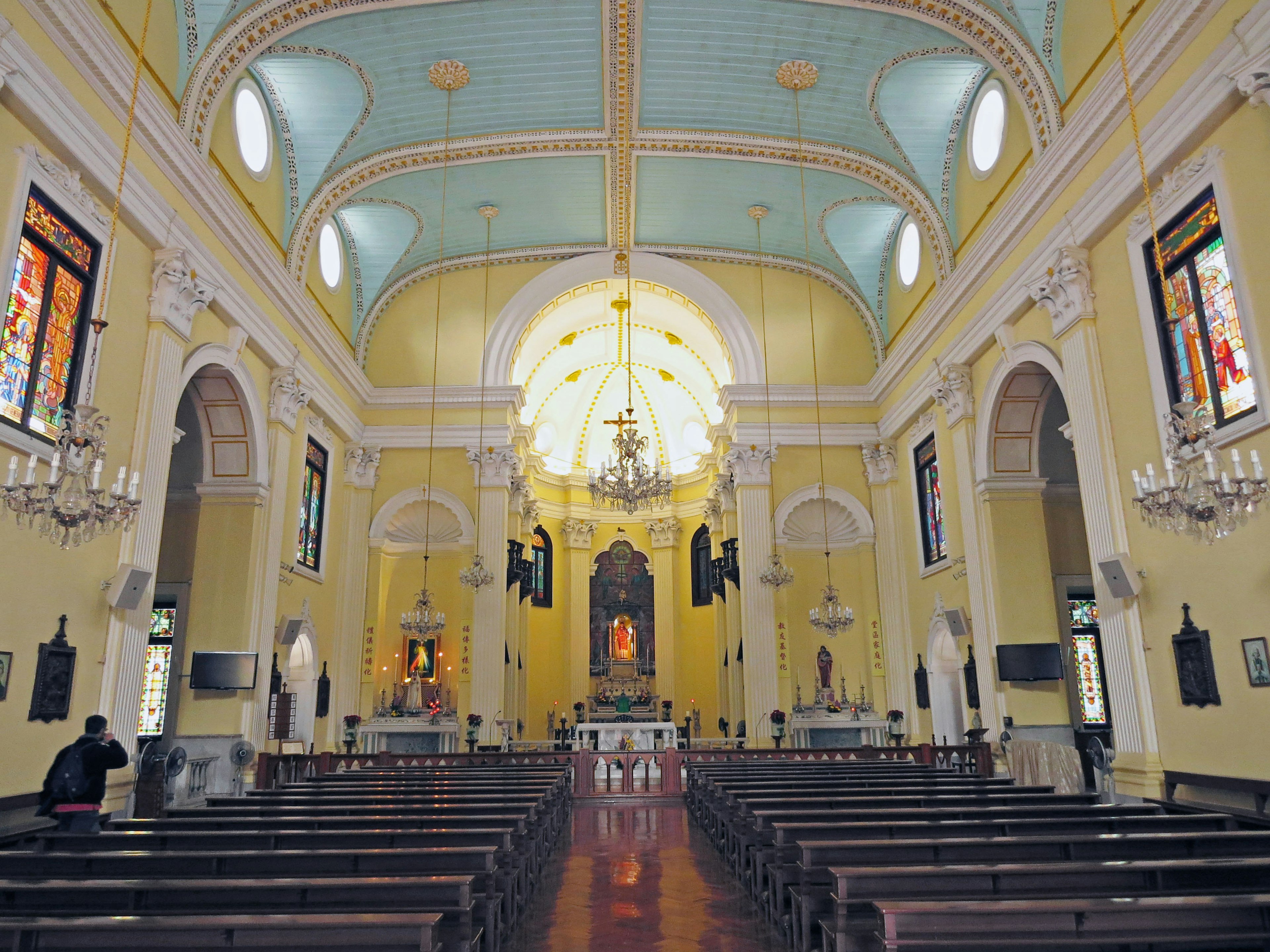 Innenraum einer Kirche mit gelben Wänden, farbigen Fenstern und einem Kronleuchter über dem Altar