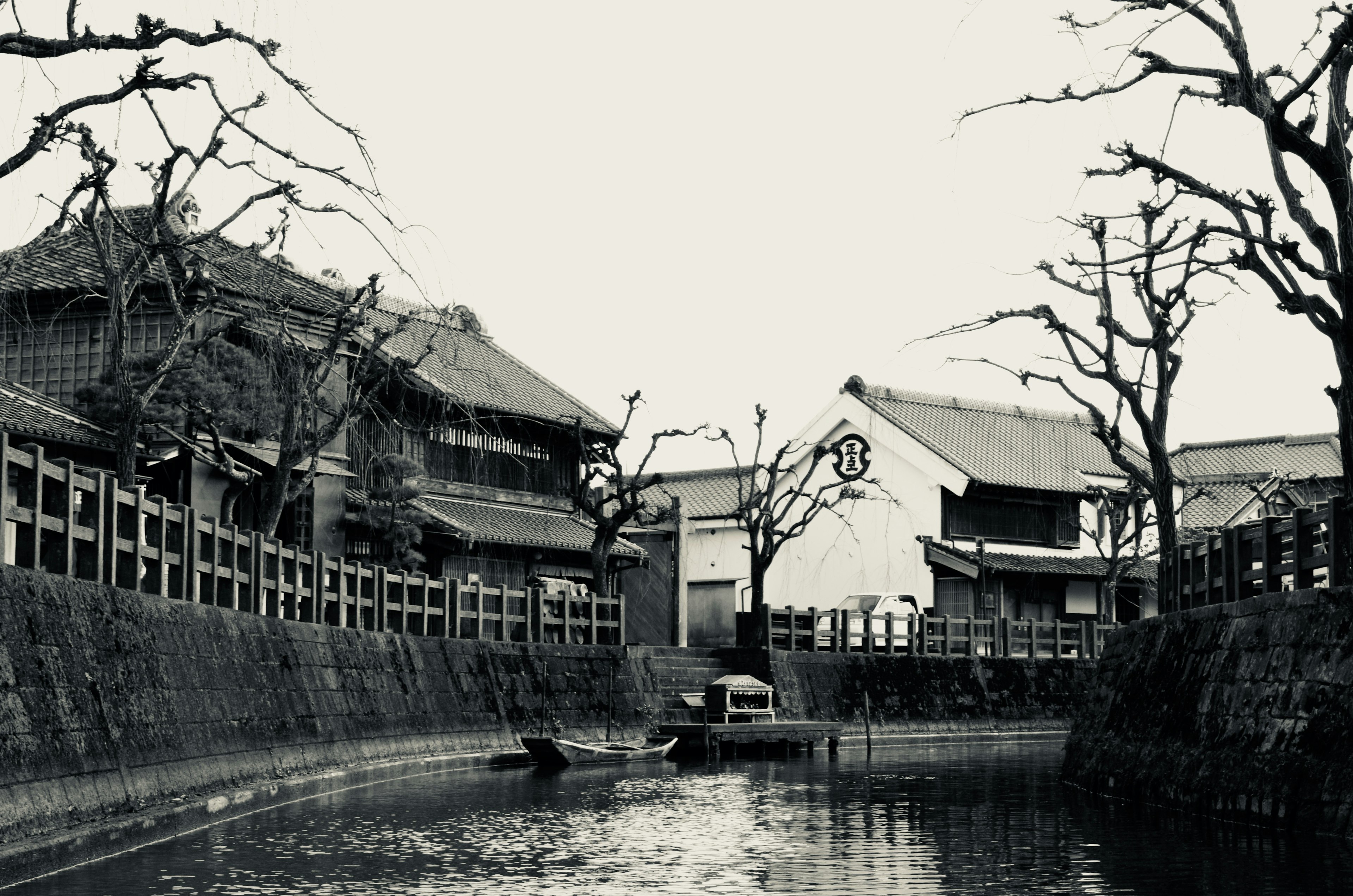 白黒の風景写真で、川沿いに並ぶ古い建物や枝のない木々が見える