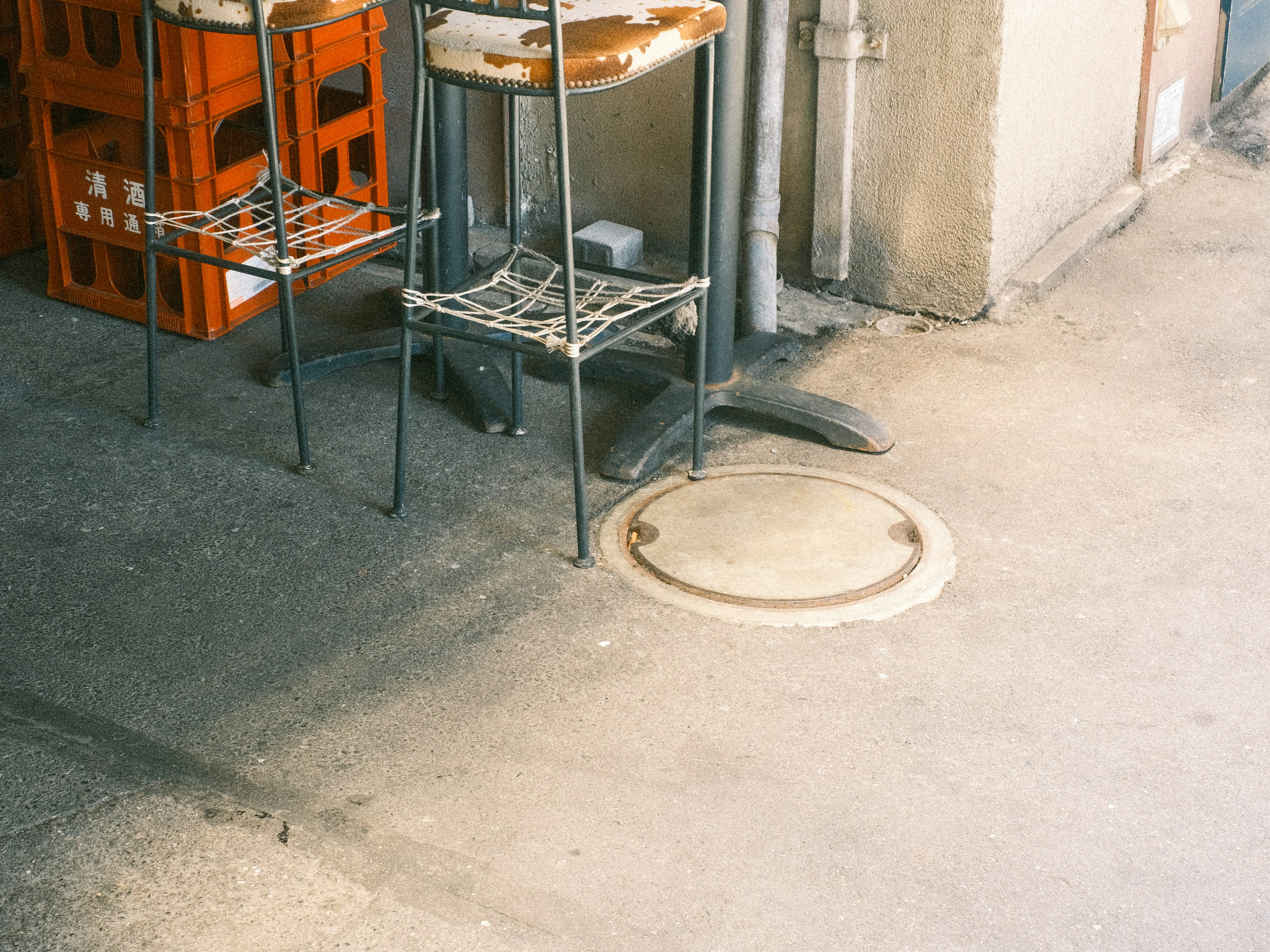 Esquina de un café con sillas de metal y una tapa de alcantarilla visible en el suelo