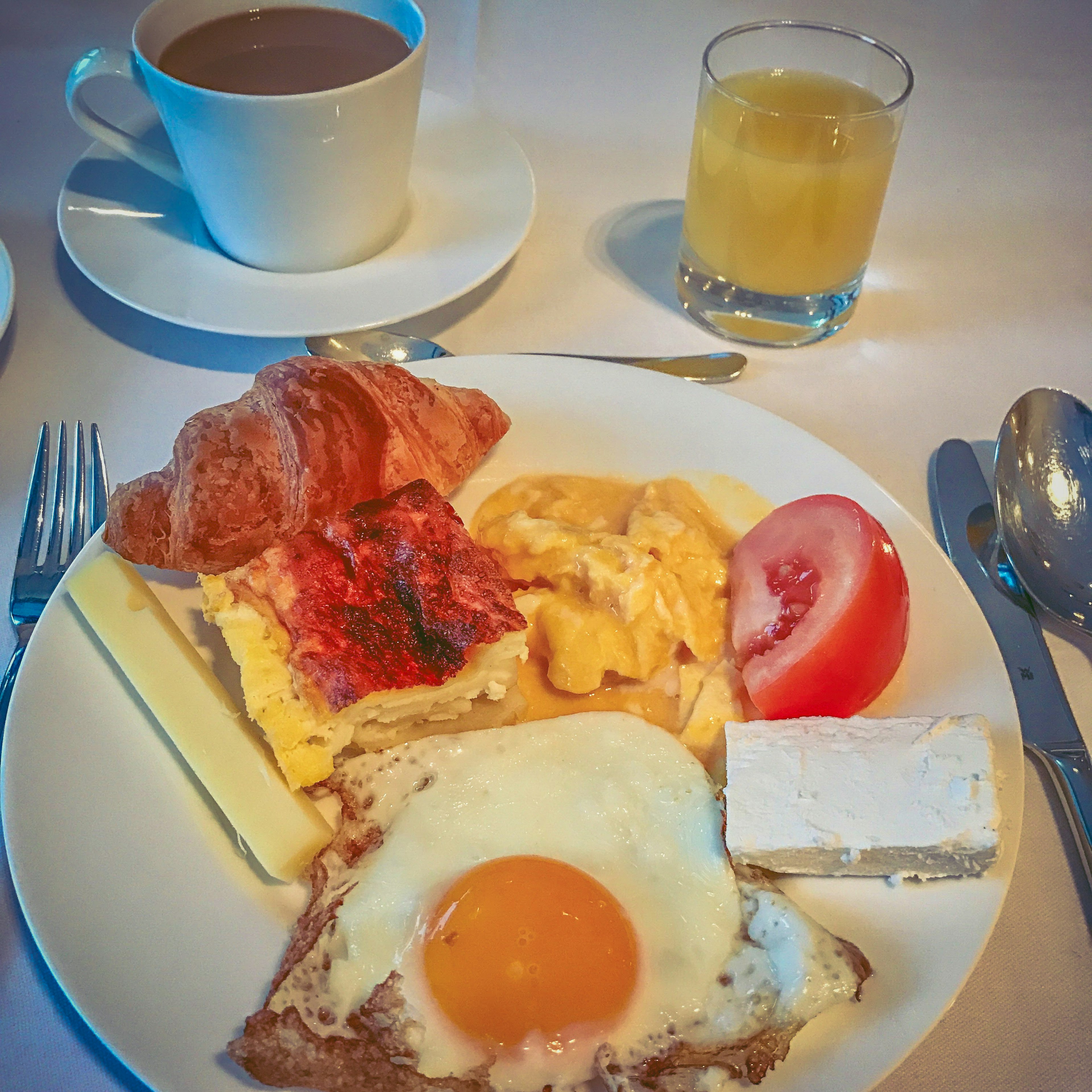 Piring sarapan dengan telur mata sapi, croissant, omelet, buah, dan minuman