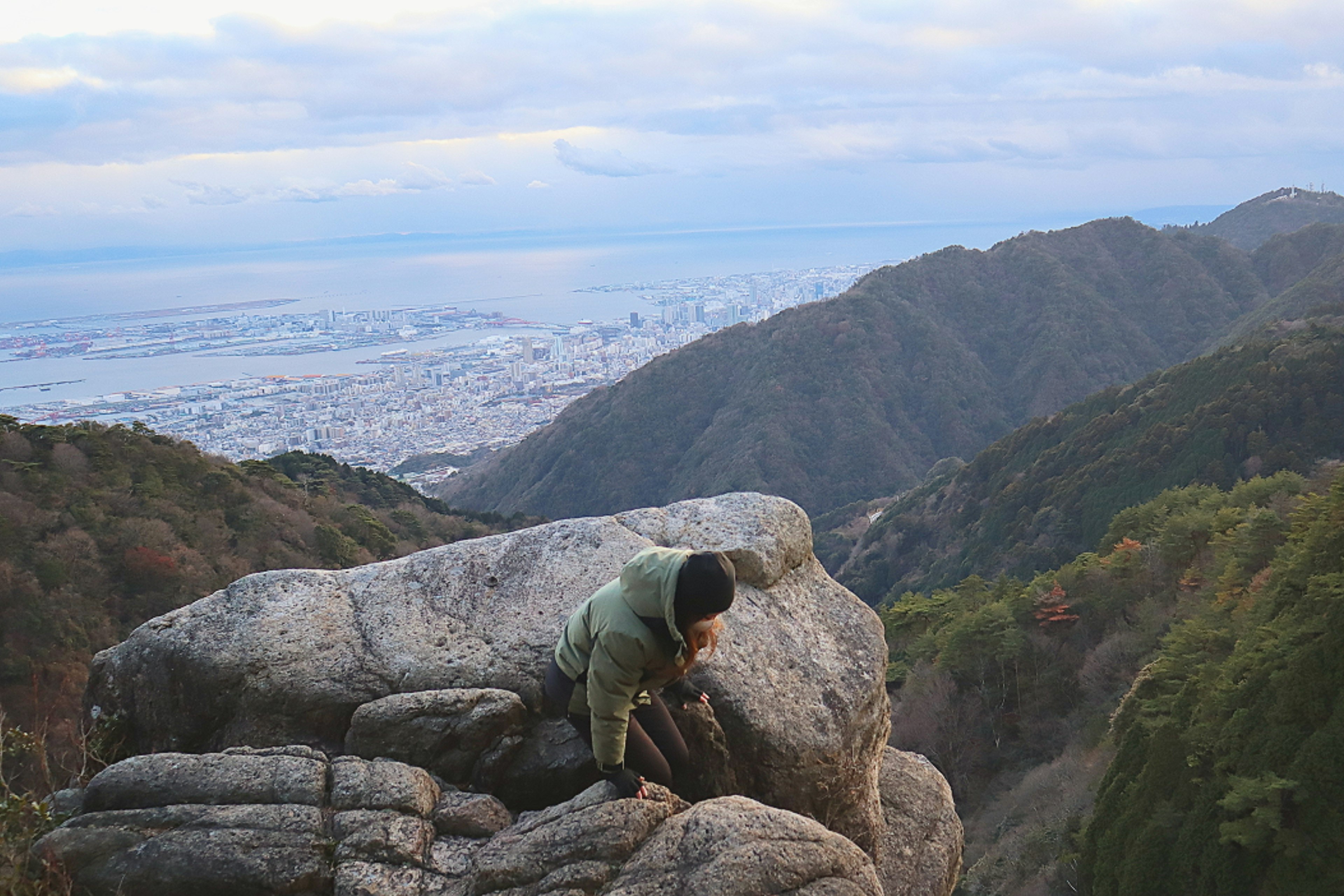 山の頂上で岩に座っている人物と遠くの都市の景色