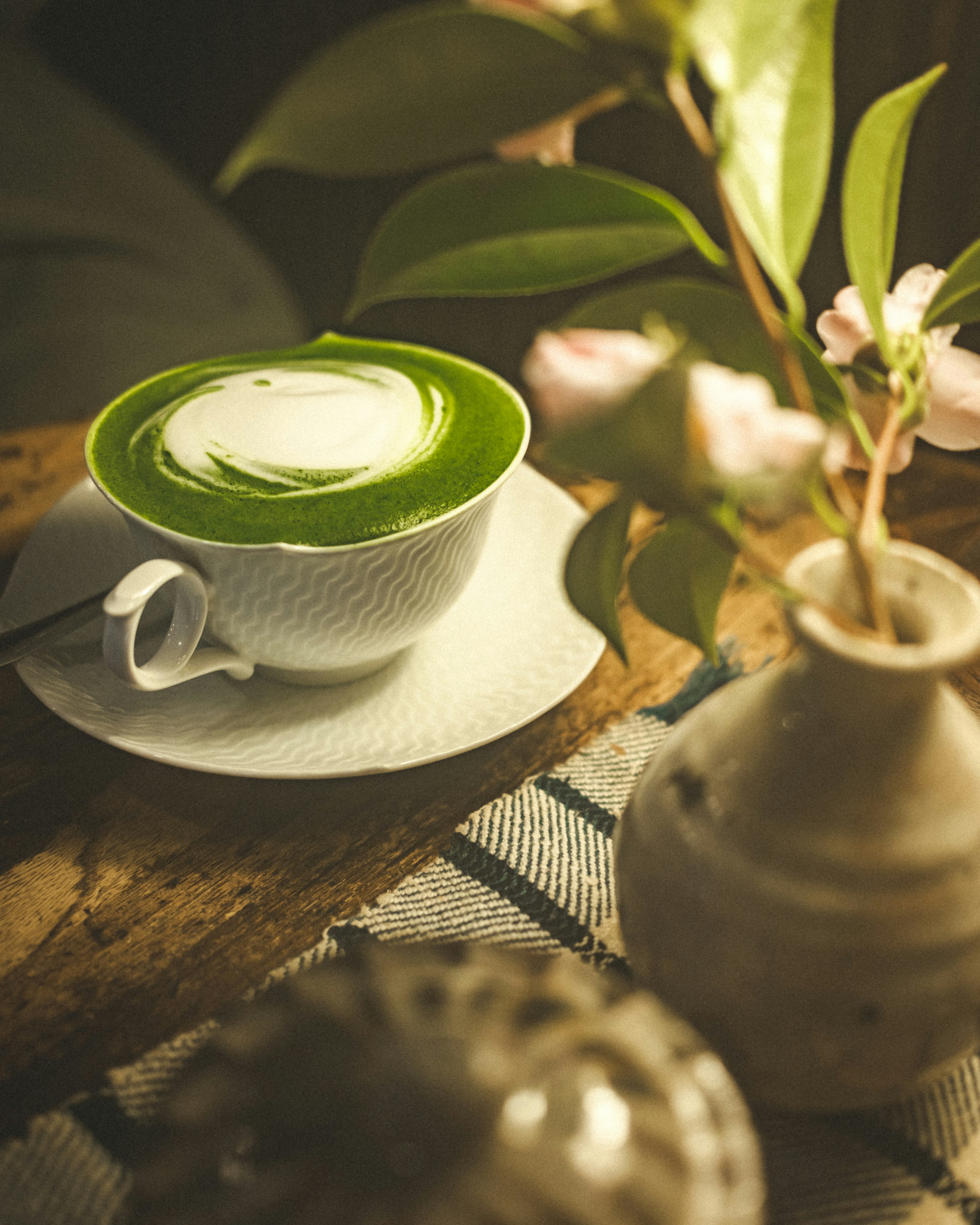 A green tea latte in a white cup with a vase of flowers on a wooden table