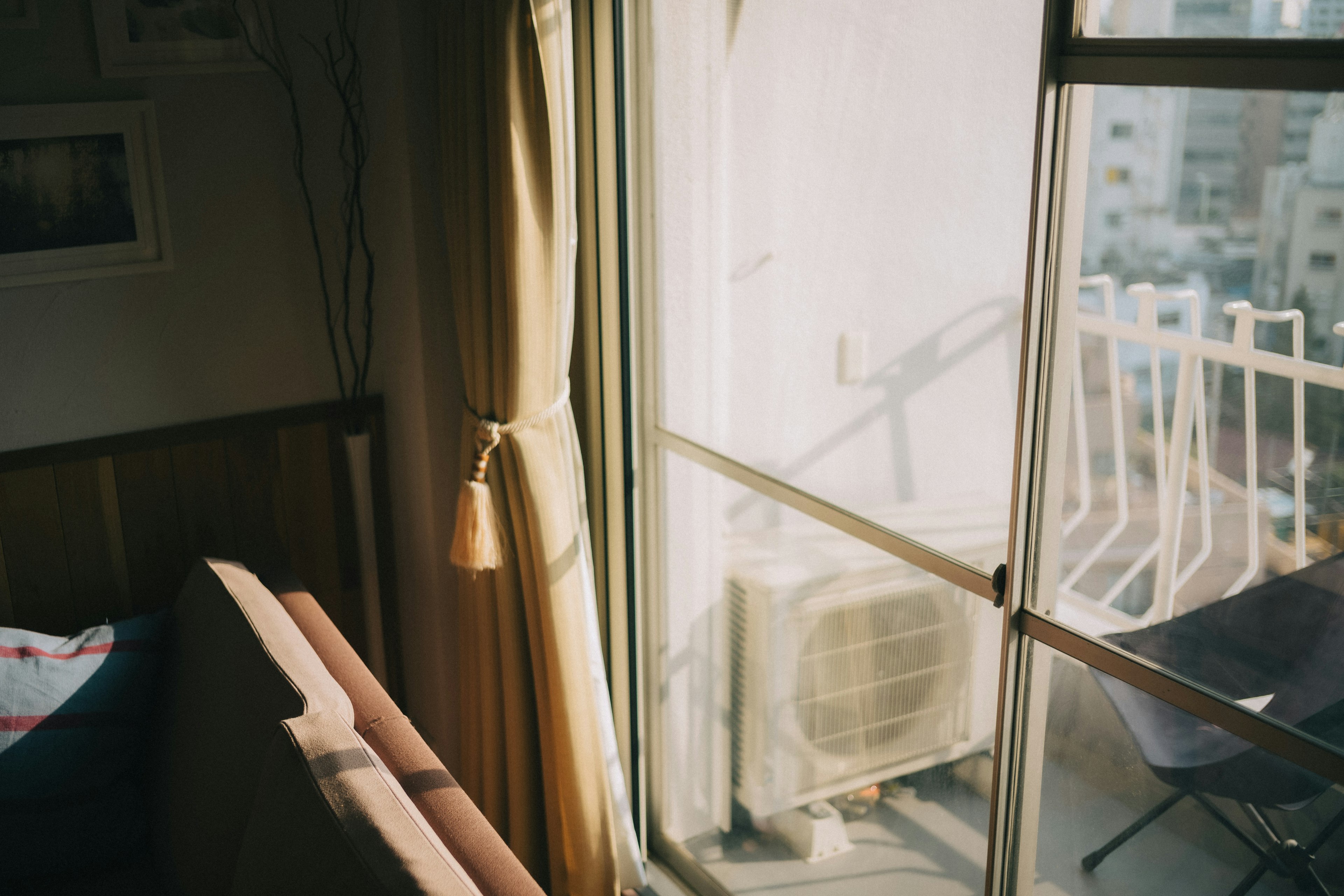 Interior de una habitación con un sofá y cortinas cerca de una ventana con un balcón y un aire acondicionado afuera