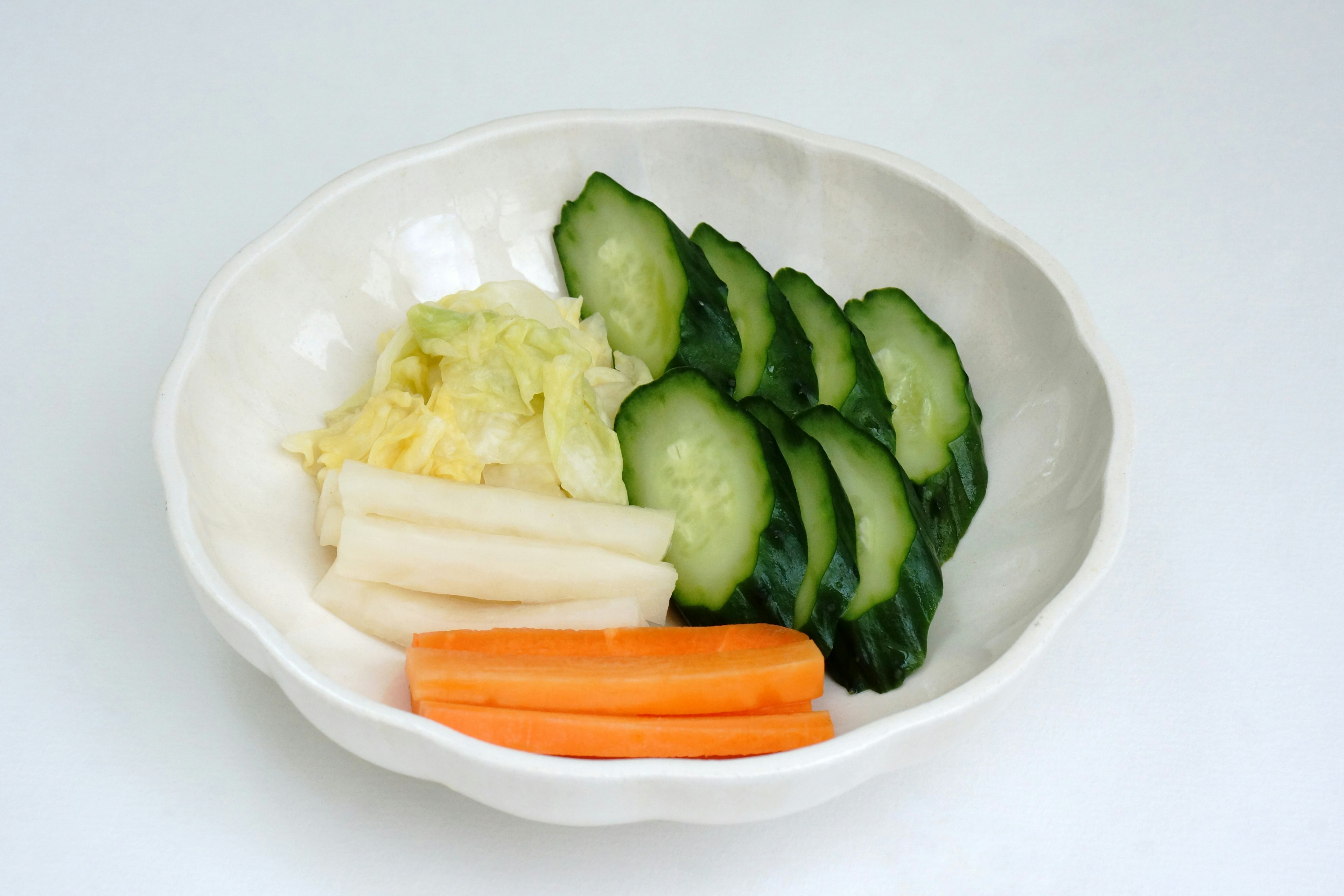 A white bowl filled with sliced cucumbers, carrots, radishes, and cabbage