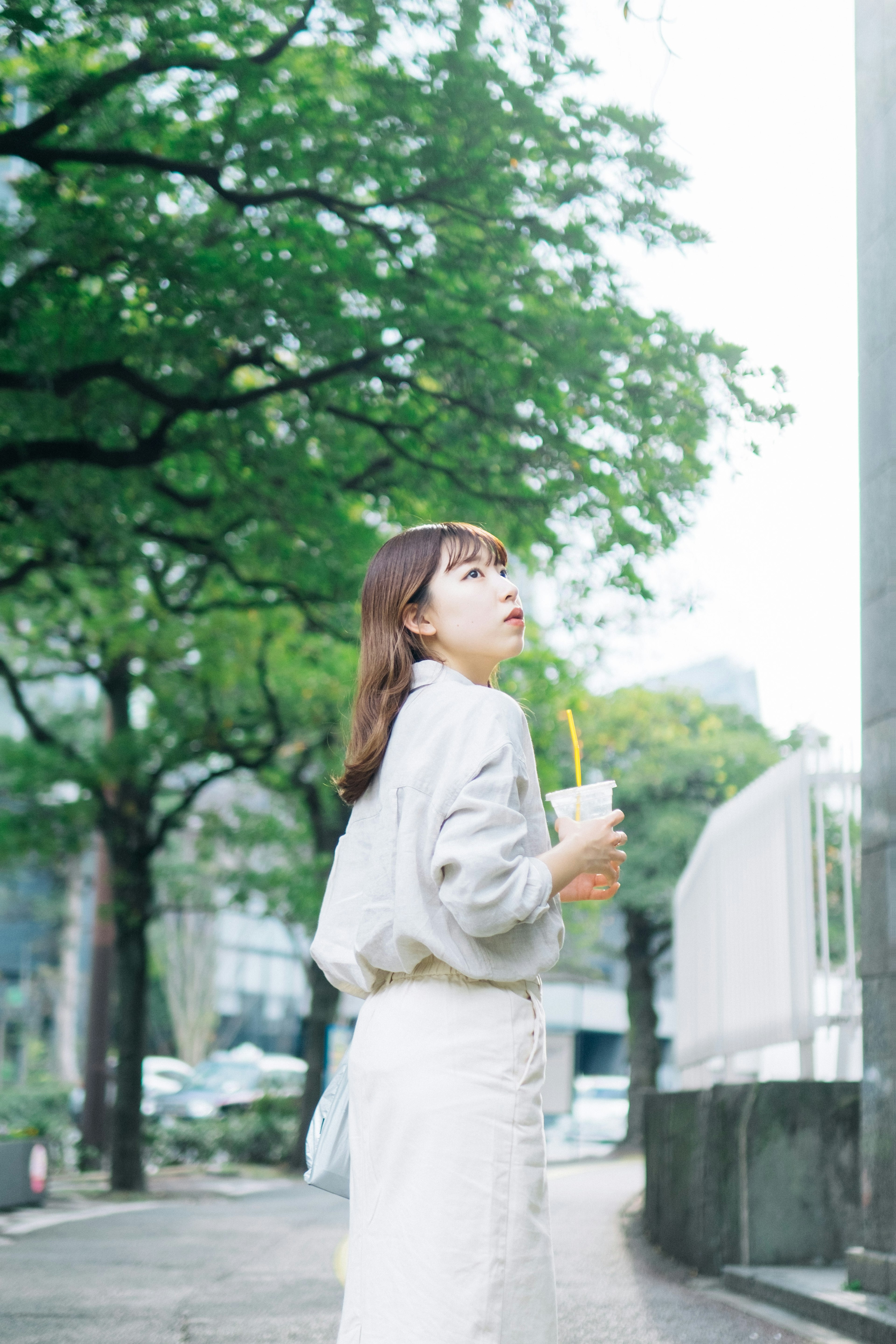 A woman holding a drink looking up at the sky among green trees