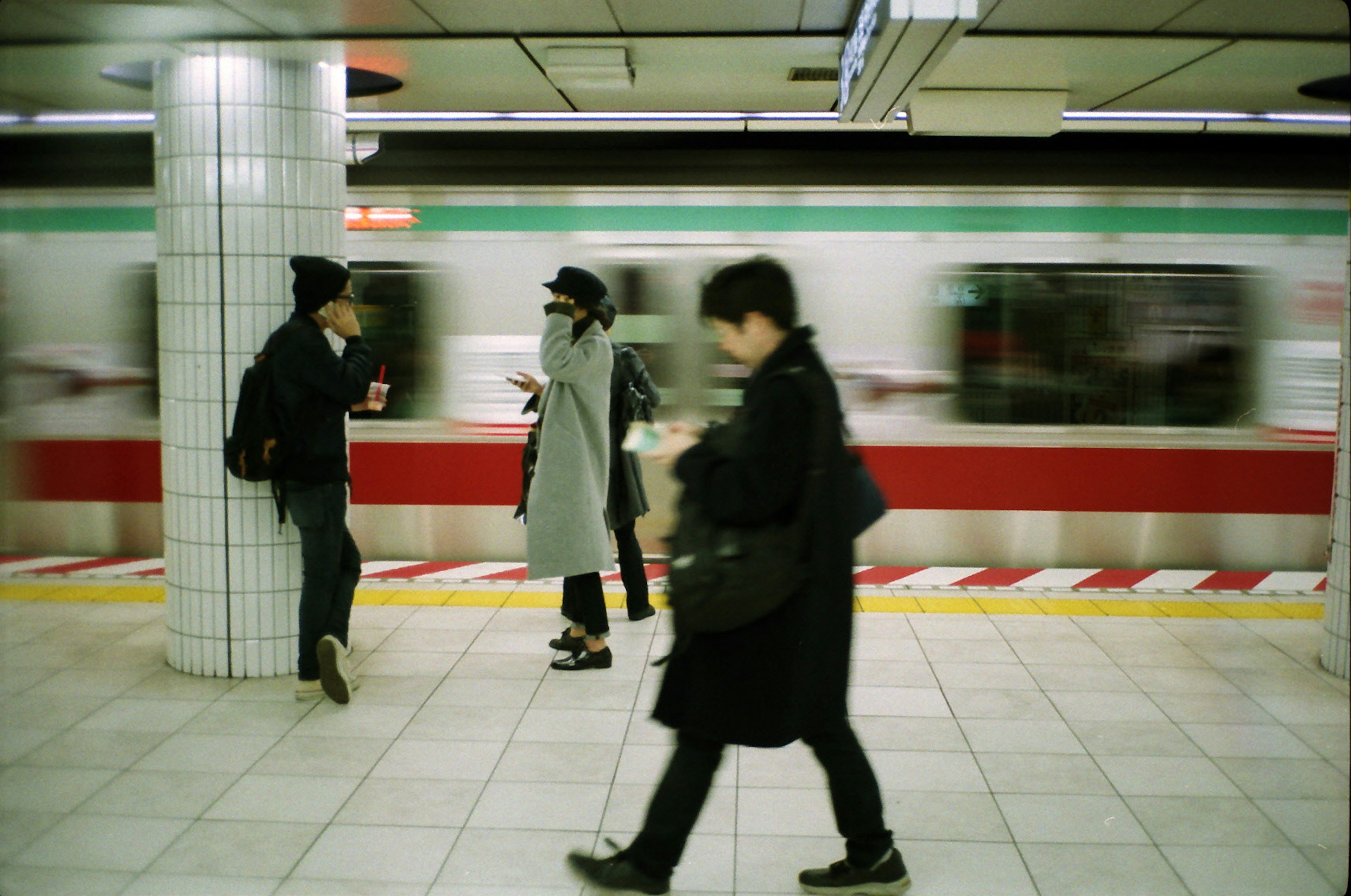 People waiting at a subway station with a moving train and white tiled column visible