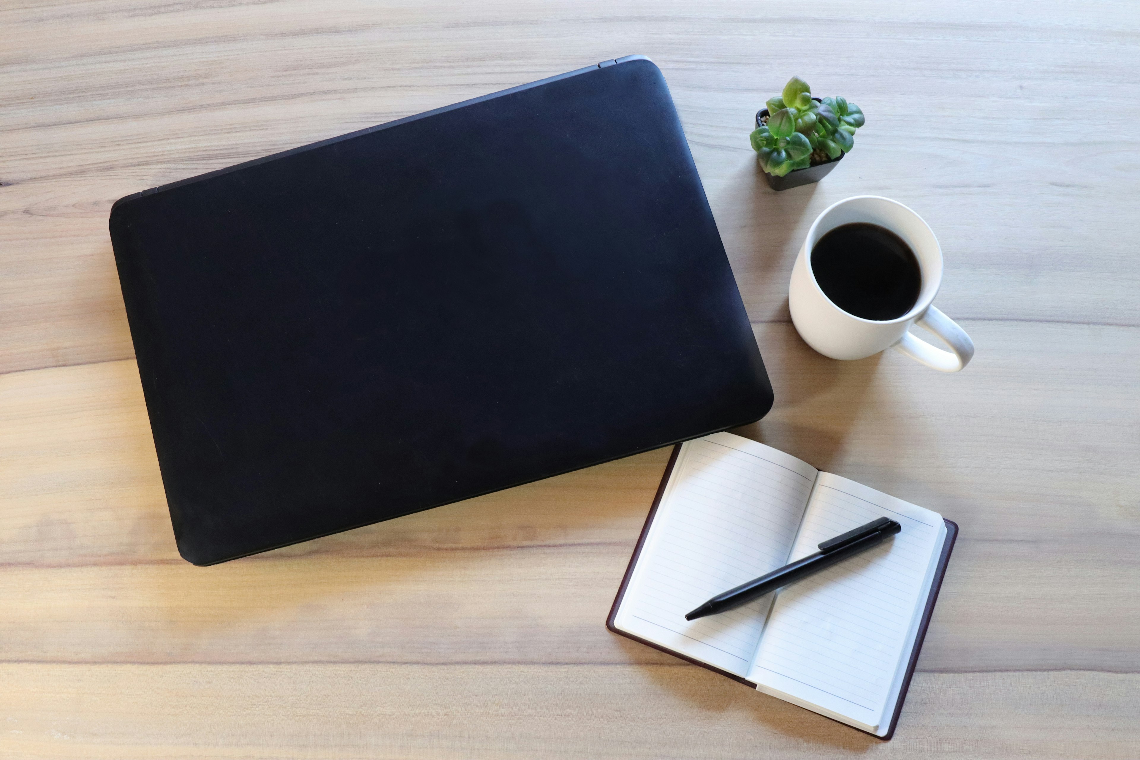 Portátil negro sobre un escritorio de madera con una taza de café blanca un cuaderno y un bolígrafo