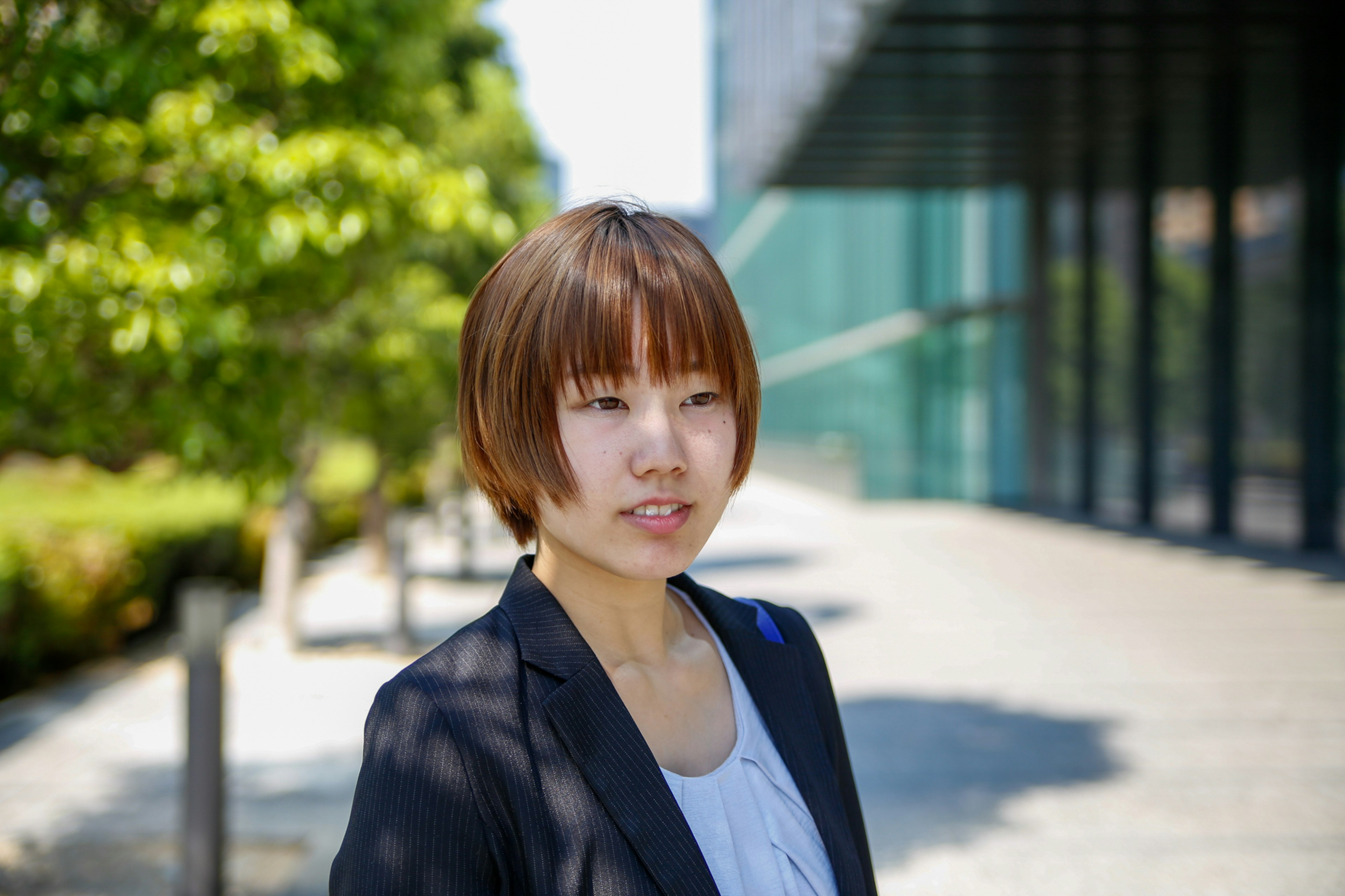 A woman in a business suit smiling outdoors with green trees and a modern building in the background