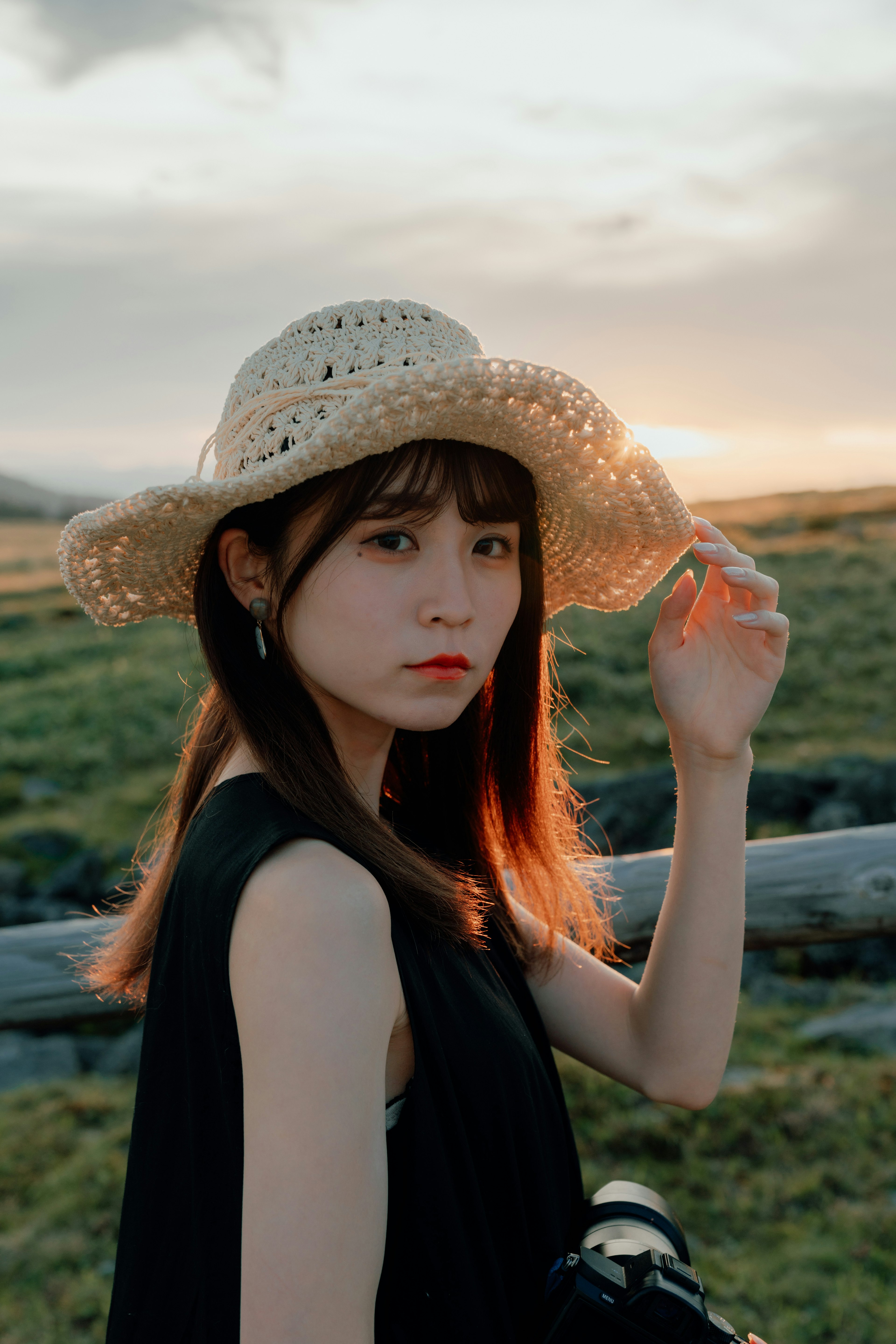 A woman wearing a straw hat raises her hand against a sunset background