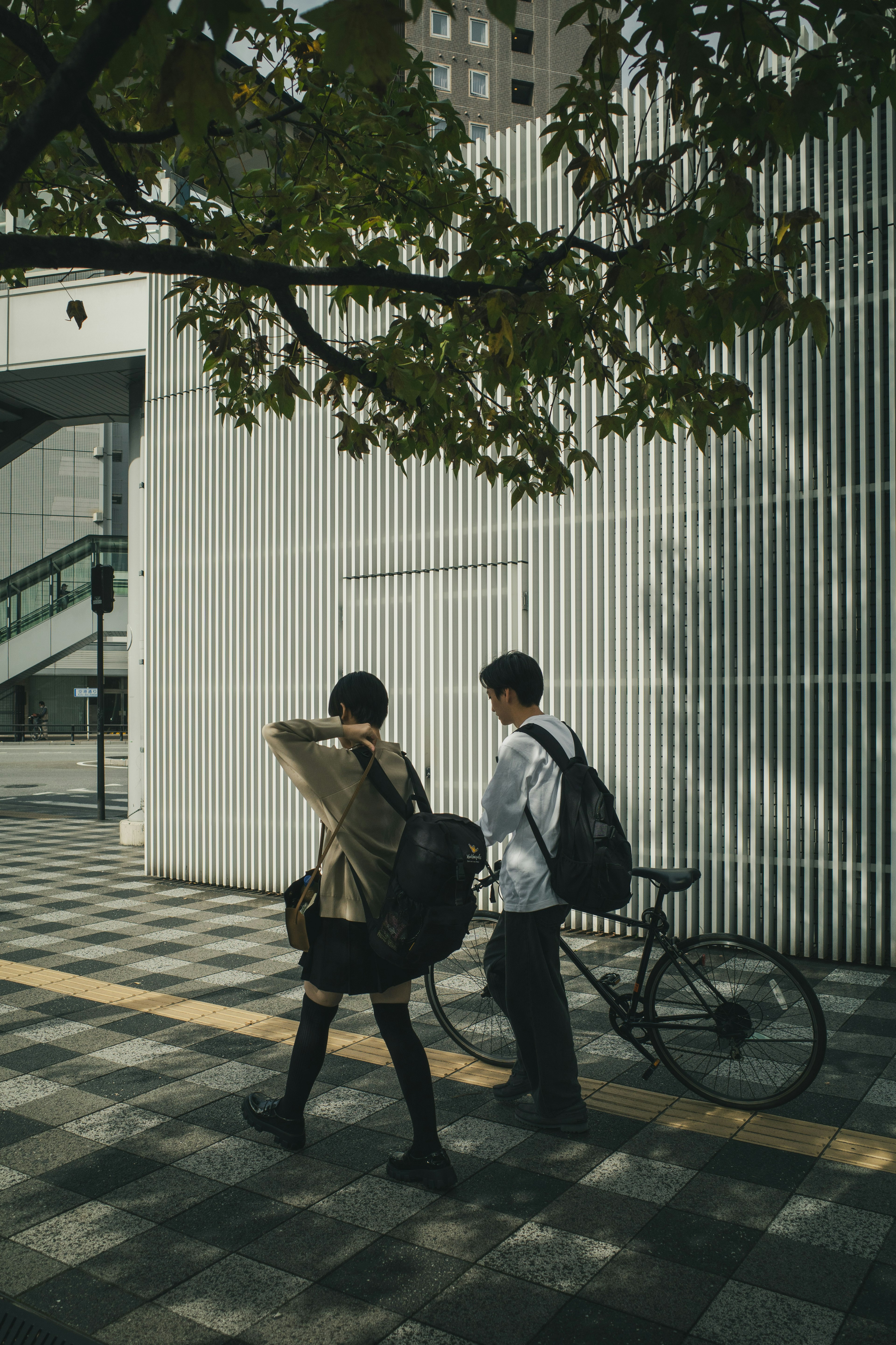 兩個人在城市人行道上走，靠近建築物和樹下的自行車