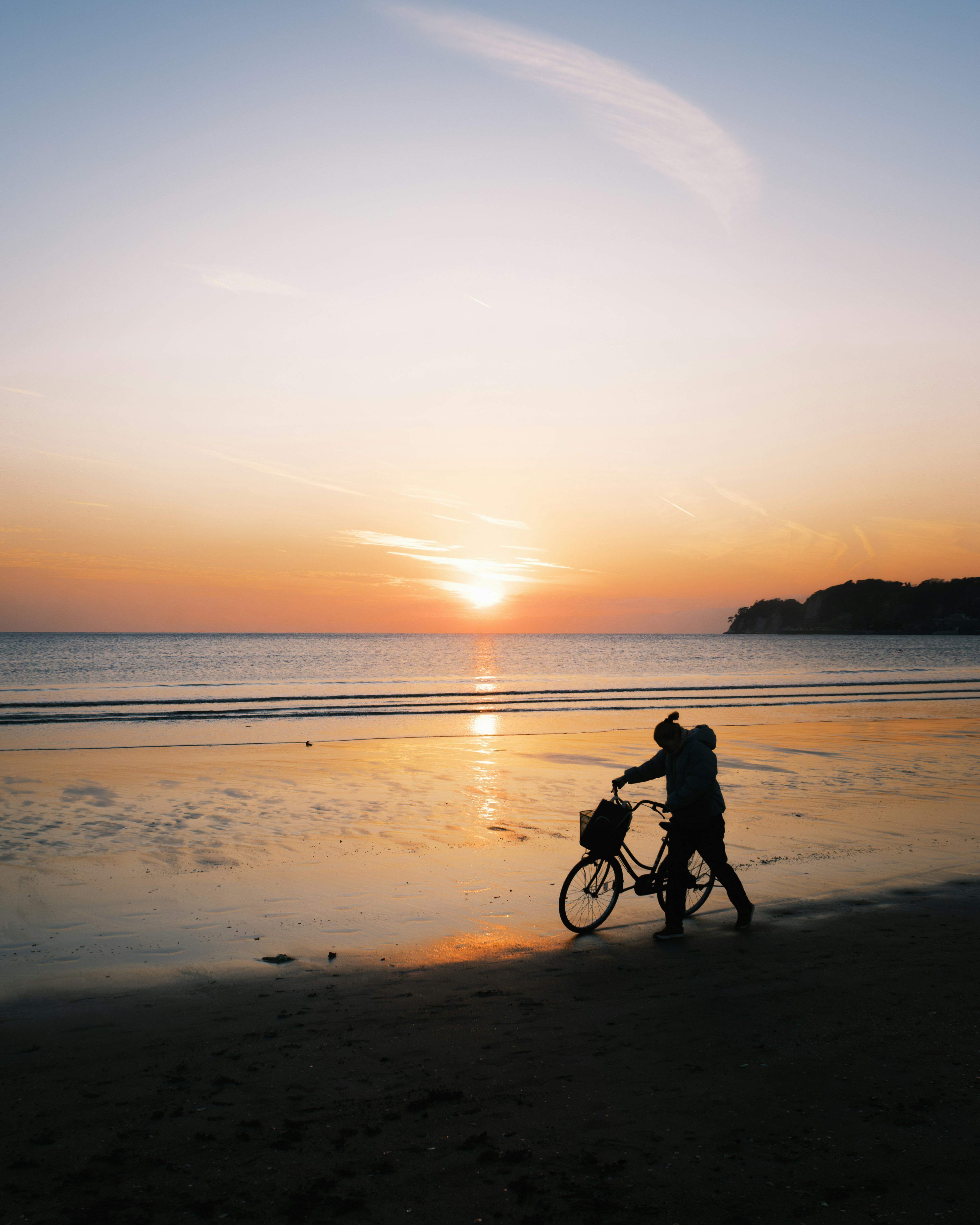 夕日を背景に自転車を押す人物が海岸を歩いている