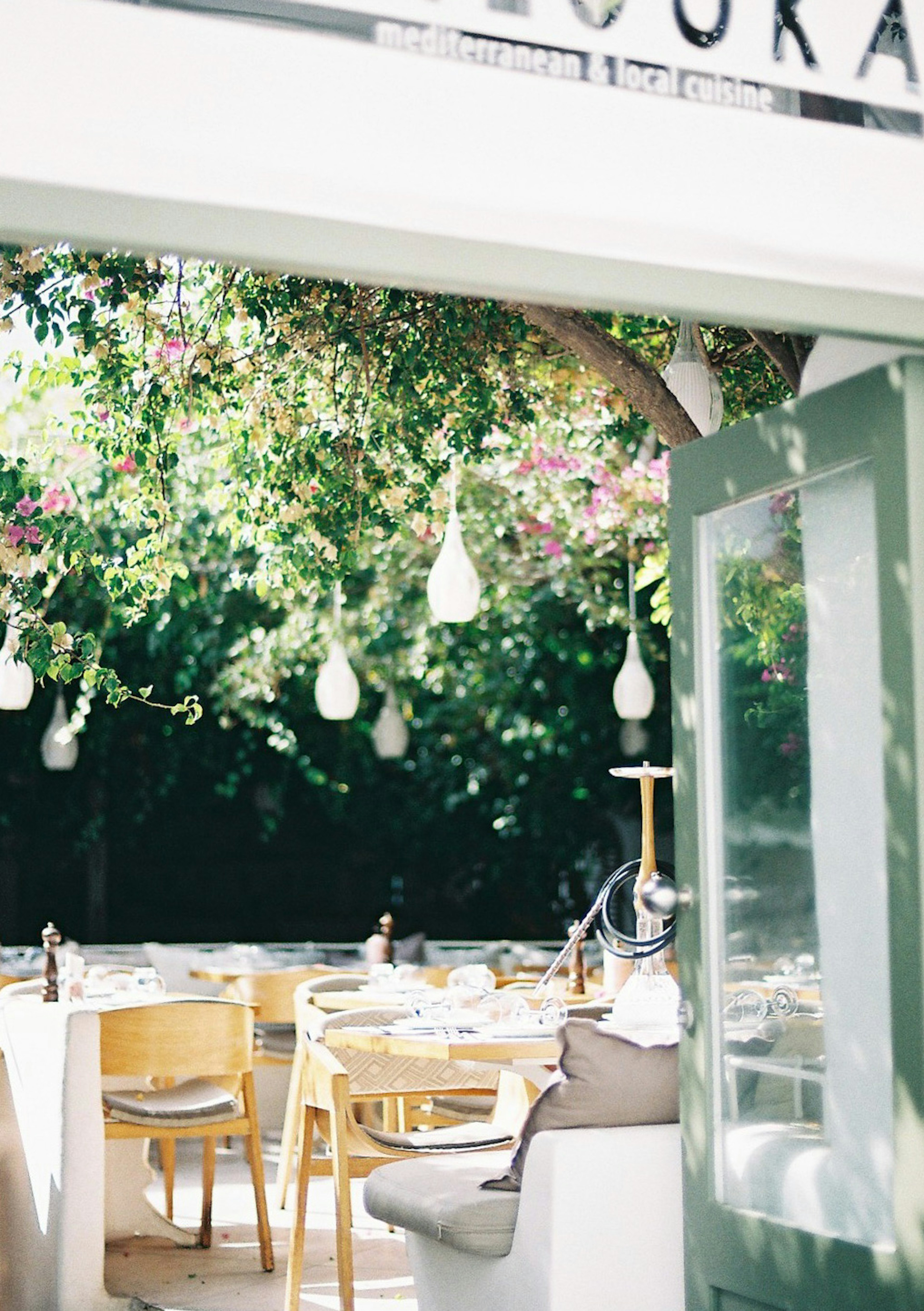 Une photo d'une terrasse de restaurant en plein air entourée de verdure avec des tables et des chaises disposées et des lumières suspendues