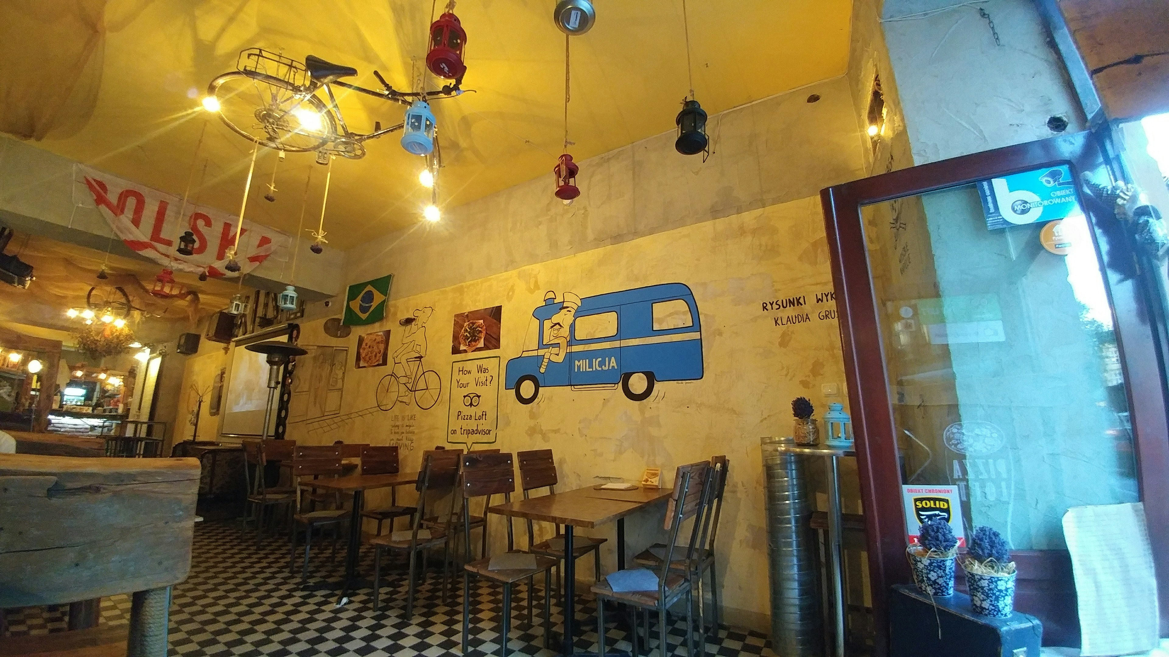 Interior de una cafetería con paredes amarillas mural de furgón azul suelo a cuadros mesas y sillas de madera