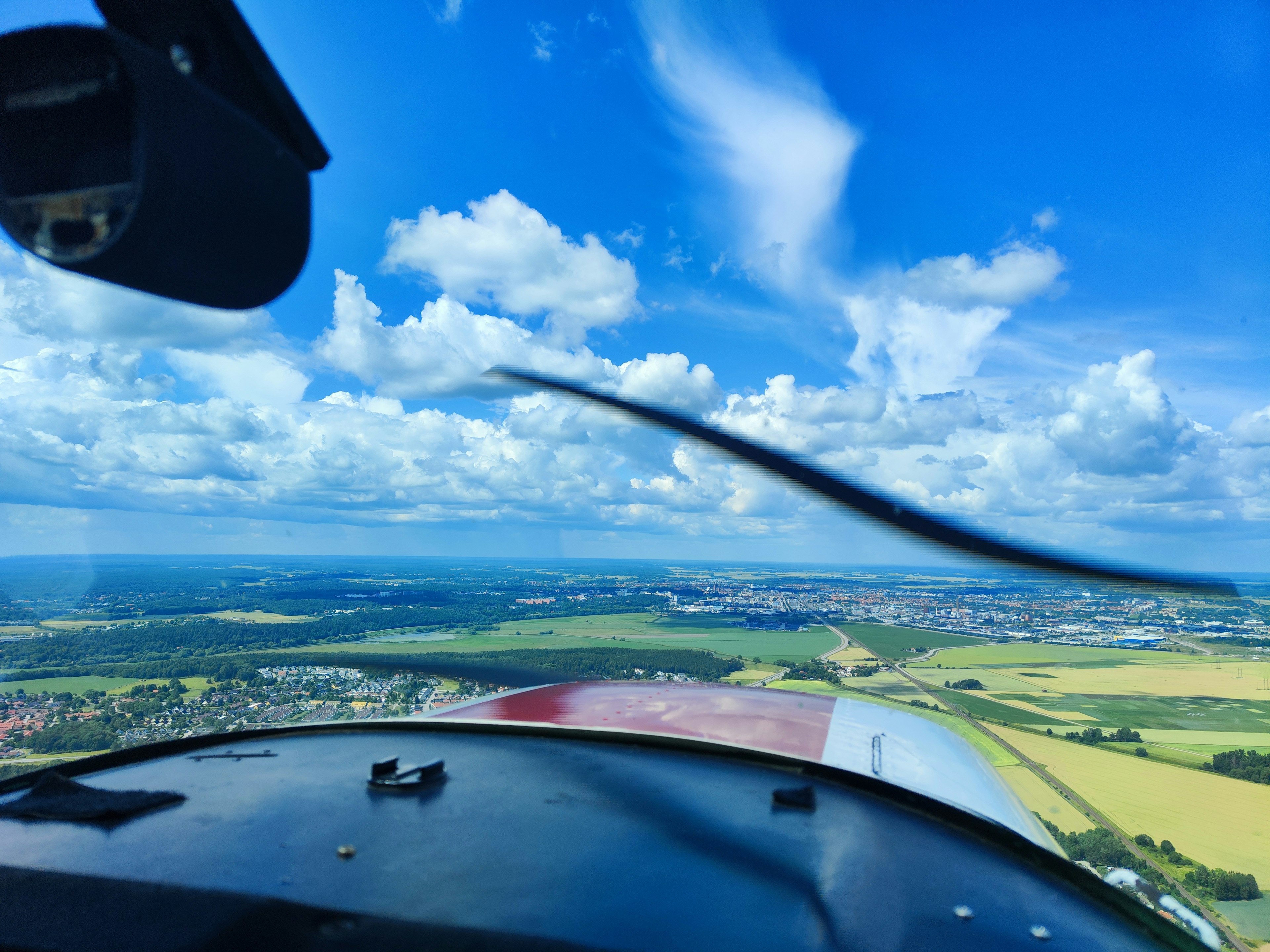 Blick aus dem Cockpit eines Flugzeugs blauer Himmel und weiße Wolken grüne Felder und eine Stadt in der Ferne