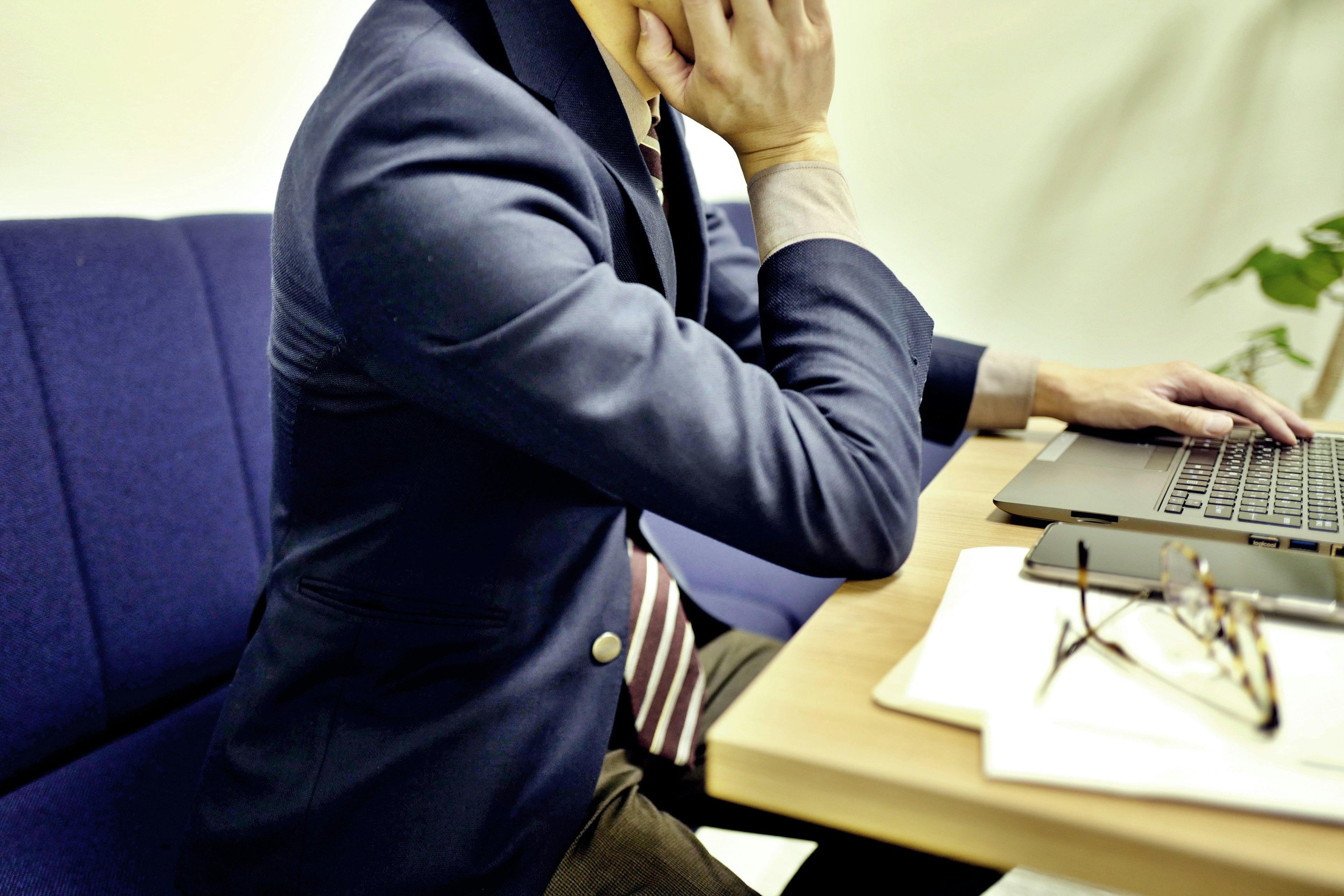 A man in a blue jacket sitting at a desk thinking while using a laptop