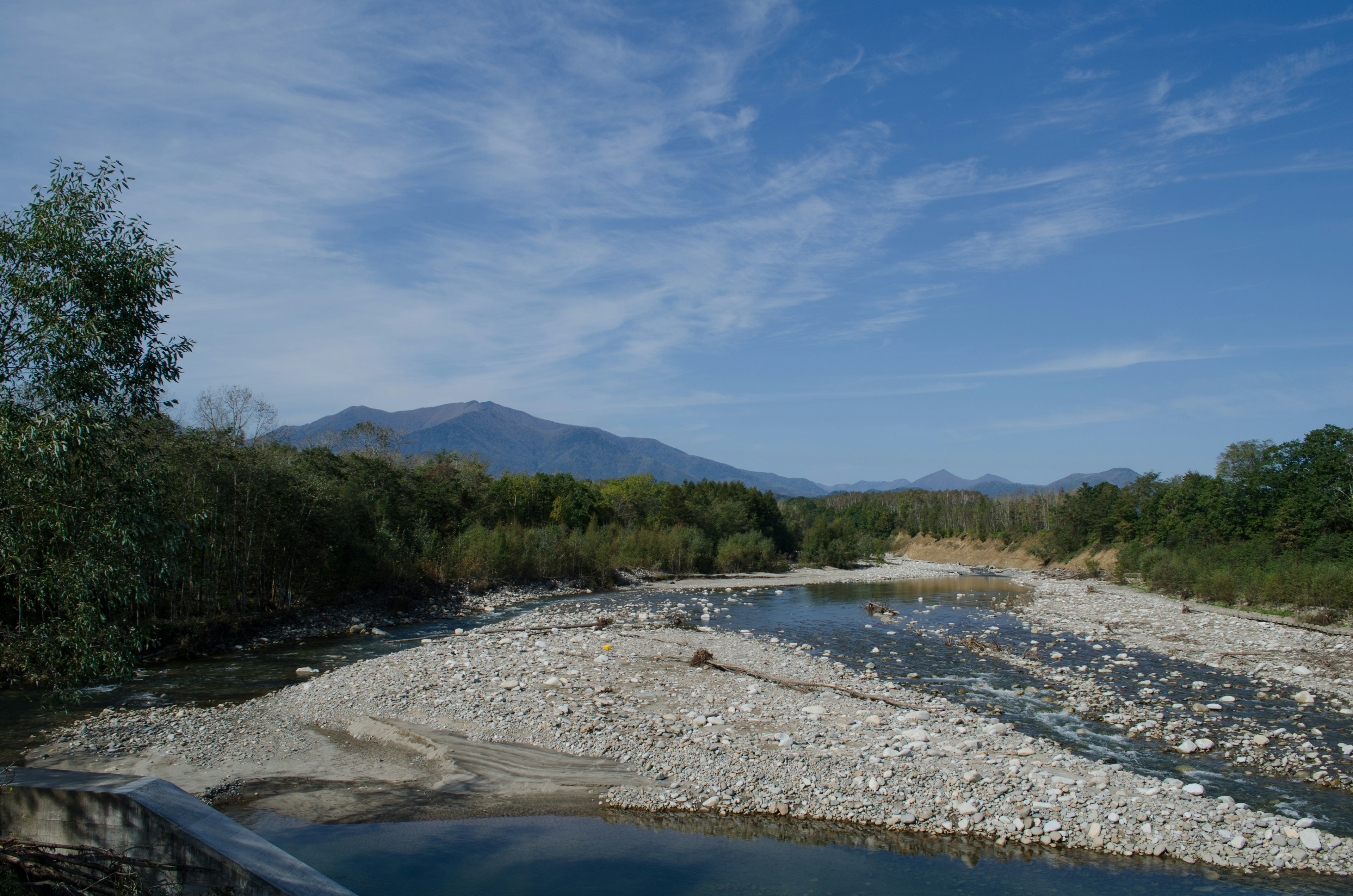 A river with a stony shore under a blue sky surrounded by green trees and distant mountains