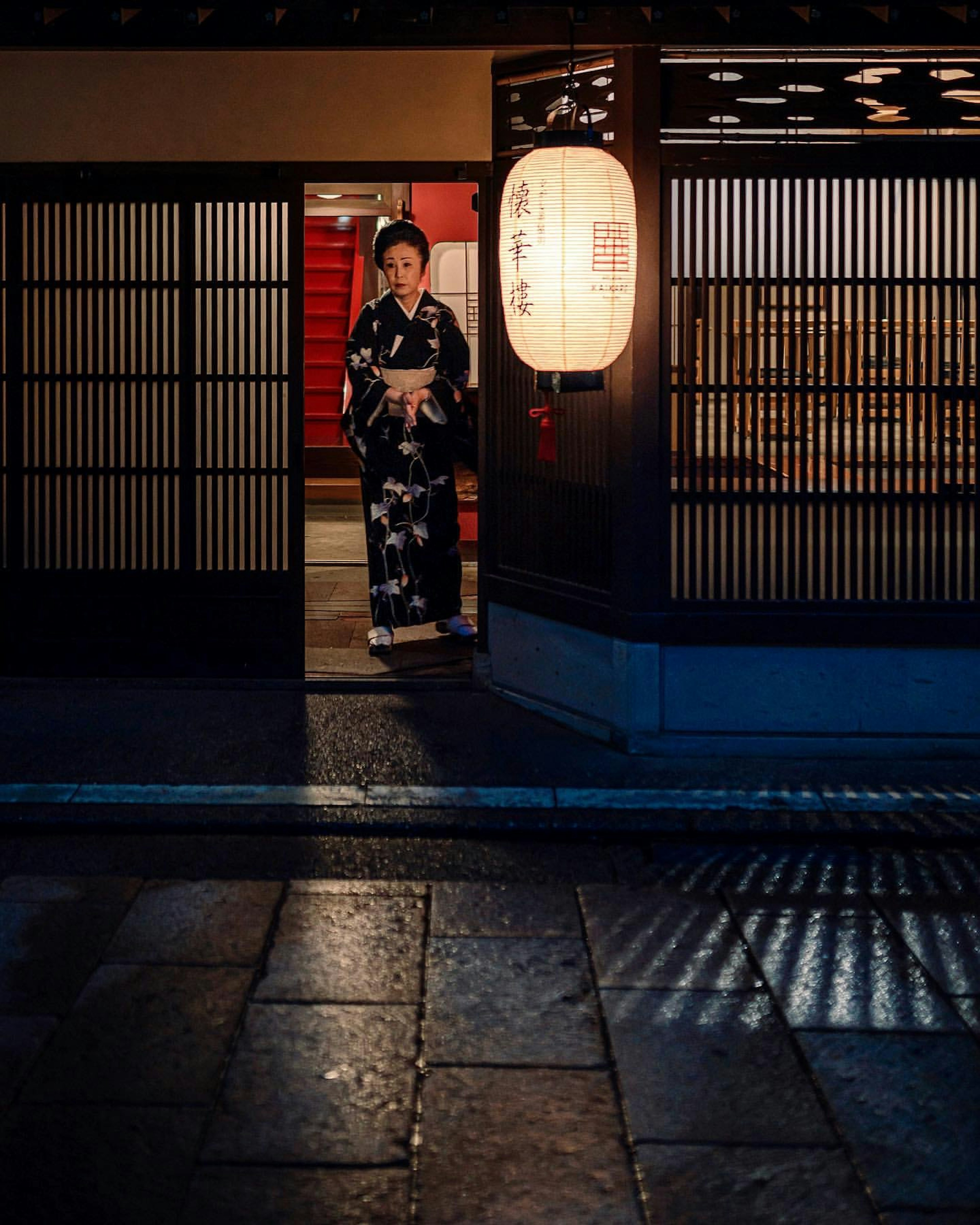 A woman in a kimono stands illuminated at night in a traditional setting