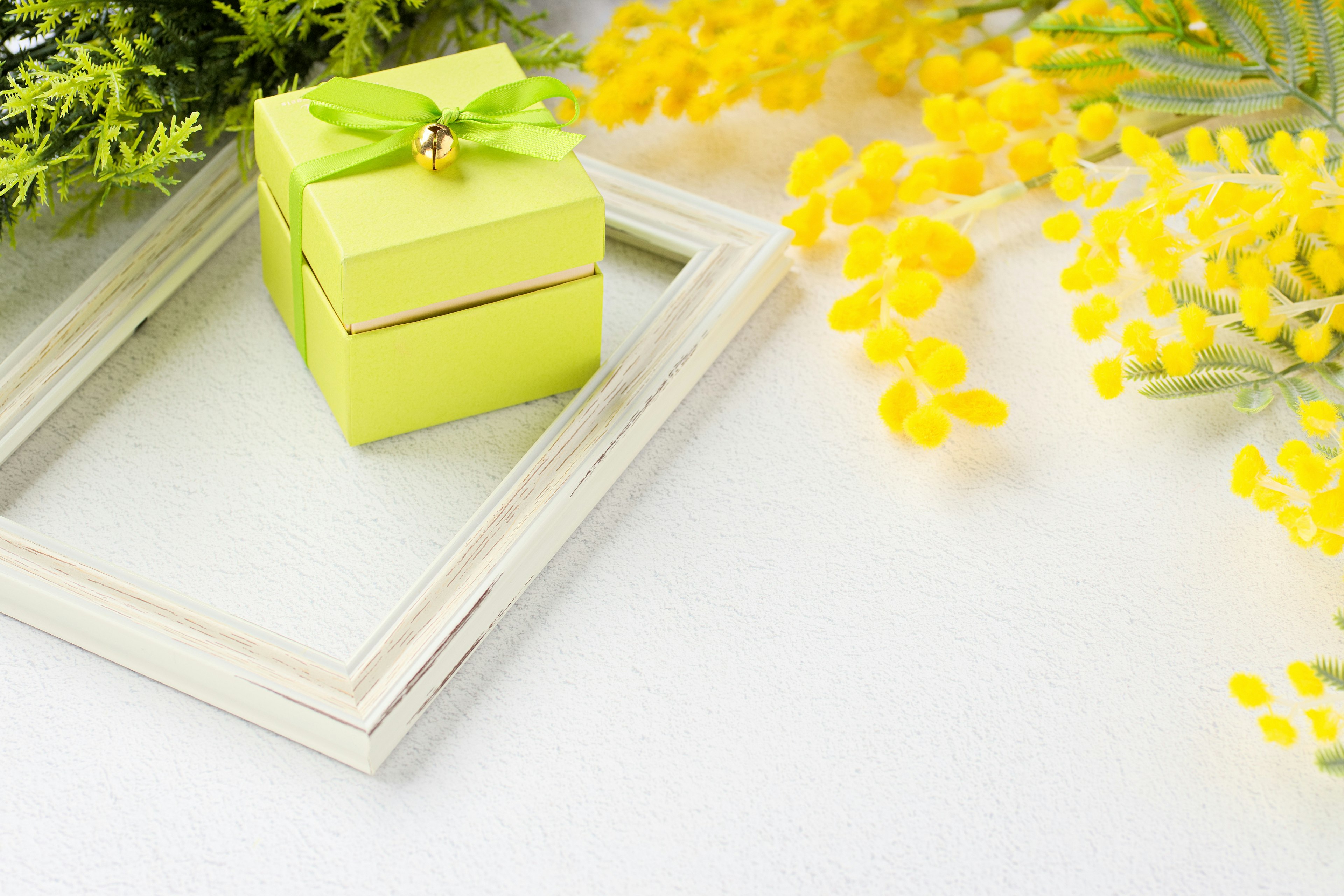 Una pequeña caja de regalo verde con un lazo colocada sobre un marco blanco rodeada de flores amarillas