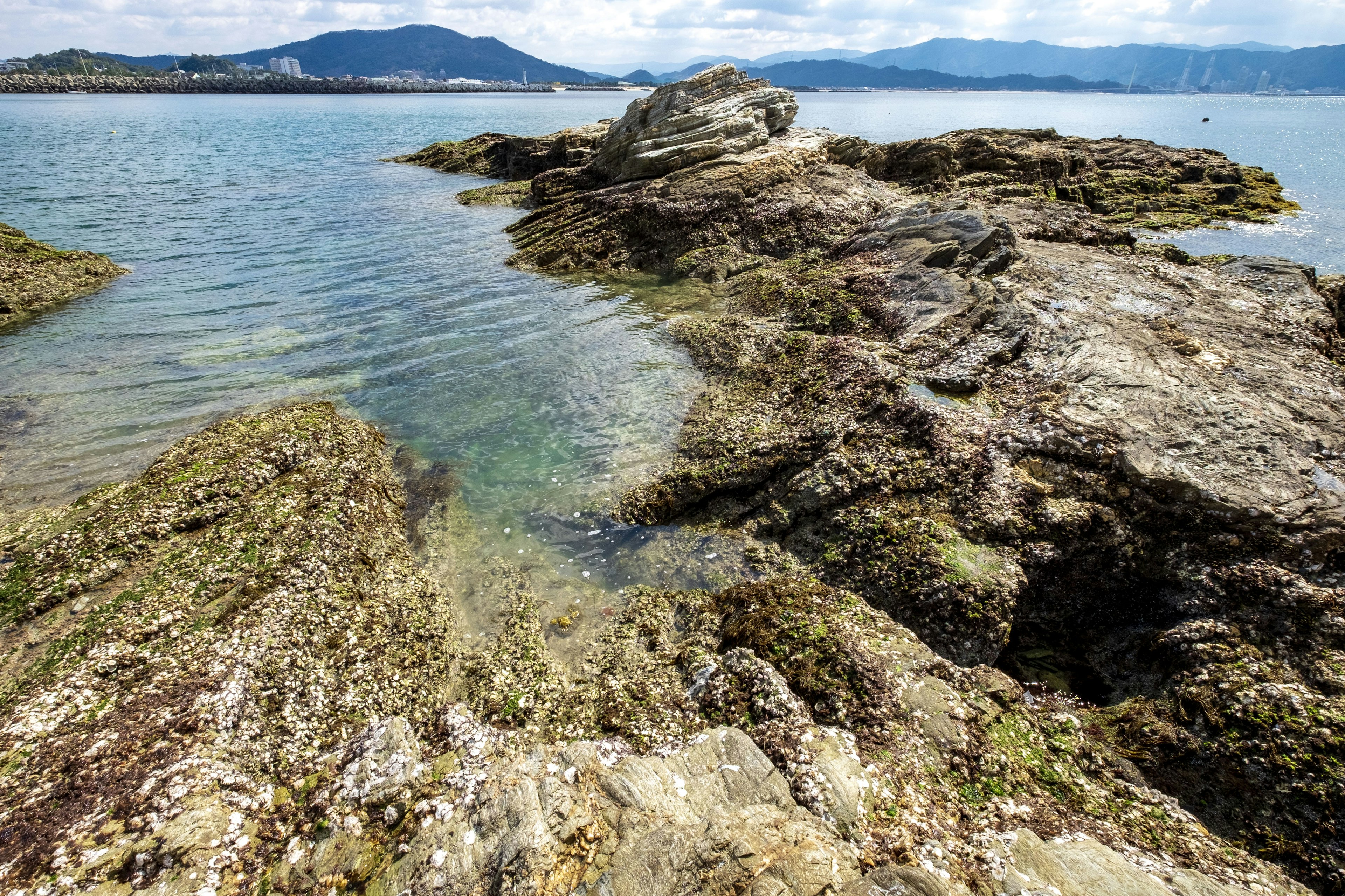 Pemandangan pantai berbatu dengan air jernih dan bukit di kejauhan