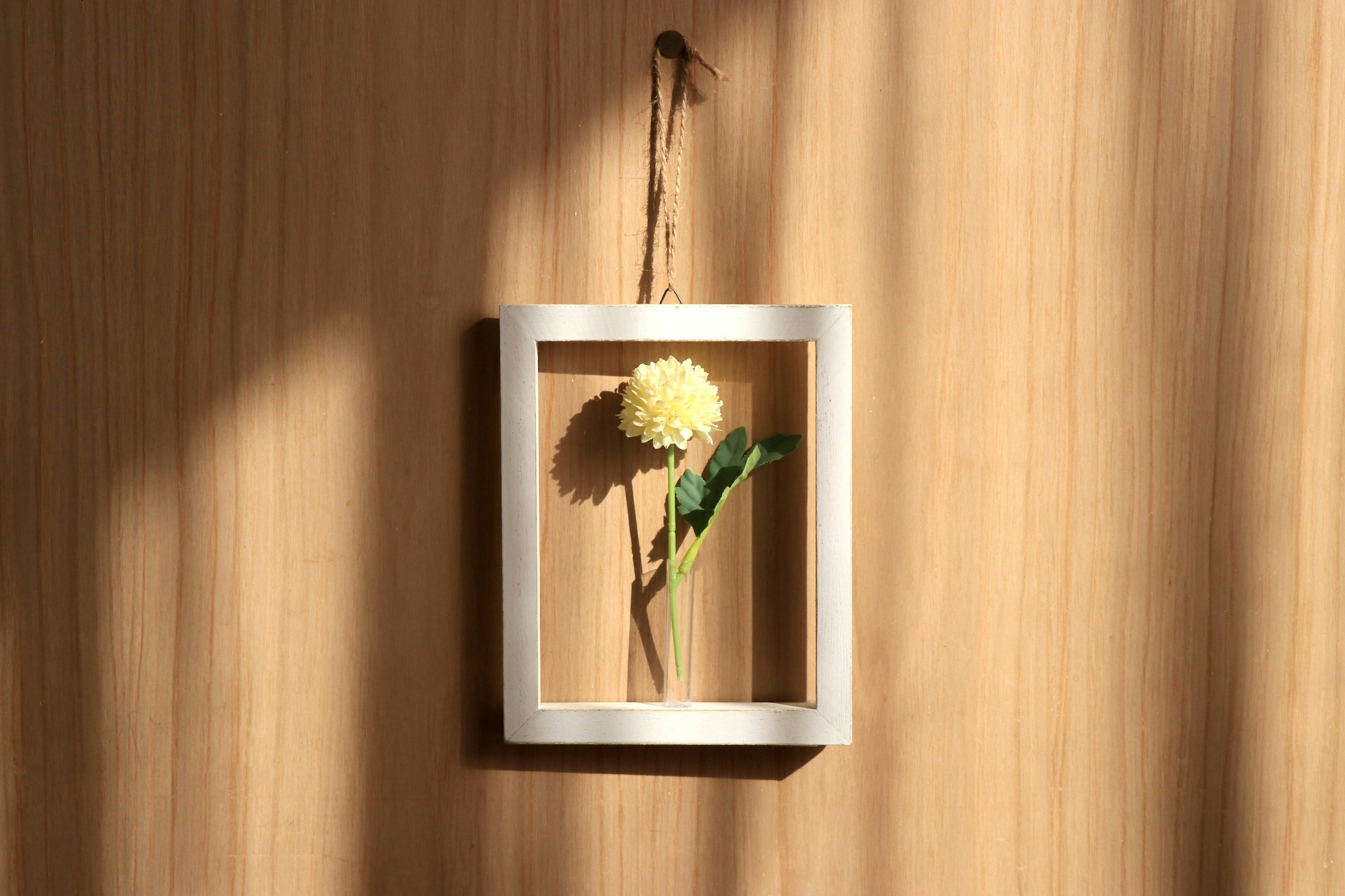 A yellow flower with green leaves displayed in a white frame hanging on a wooden wall