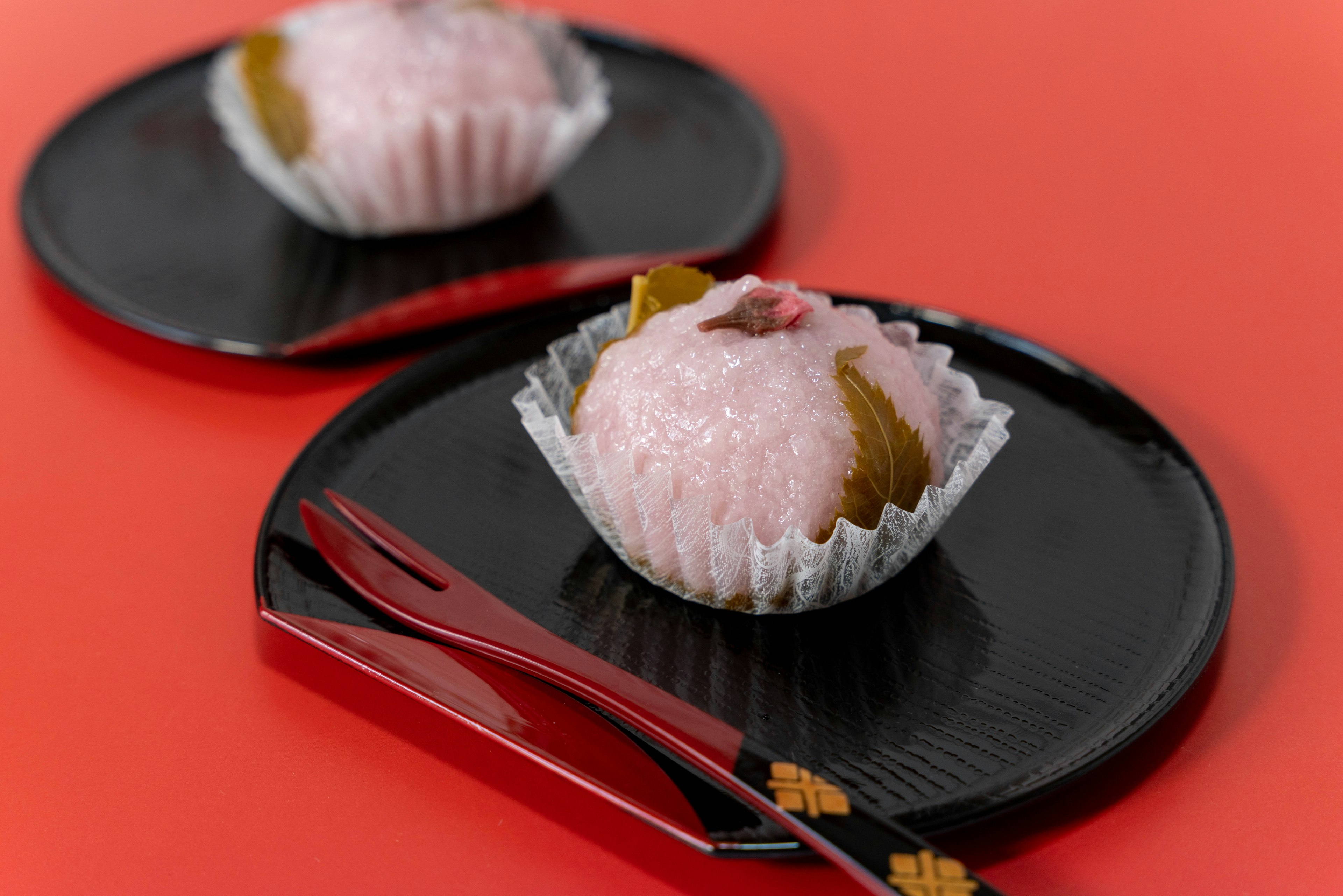 Two sakura mochi on black plates with red background and chopsticks