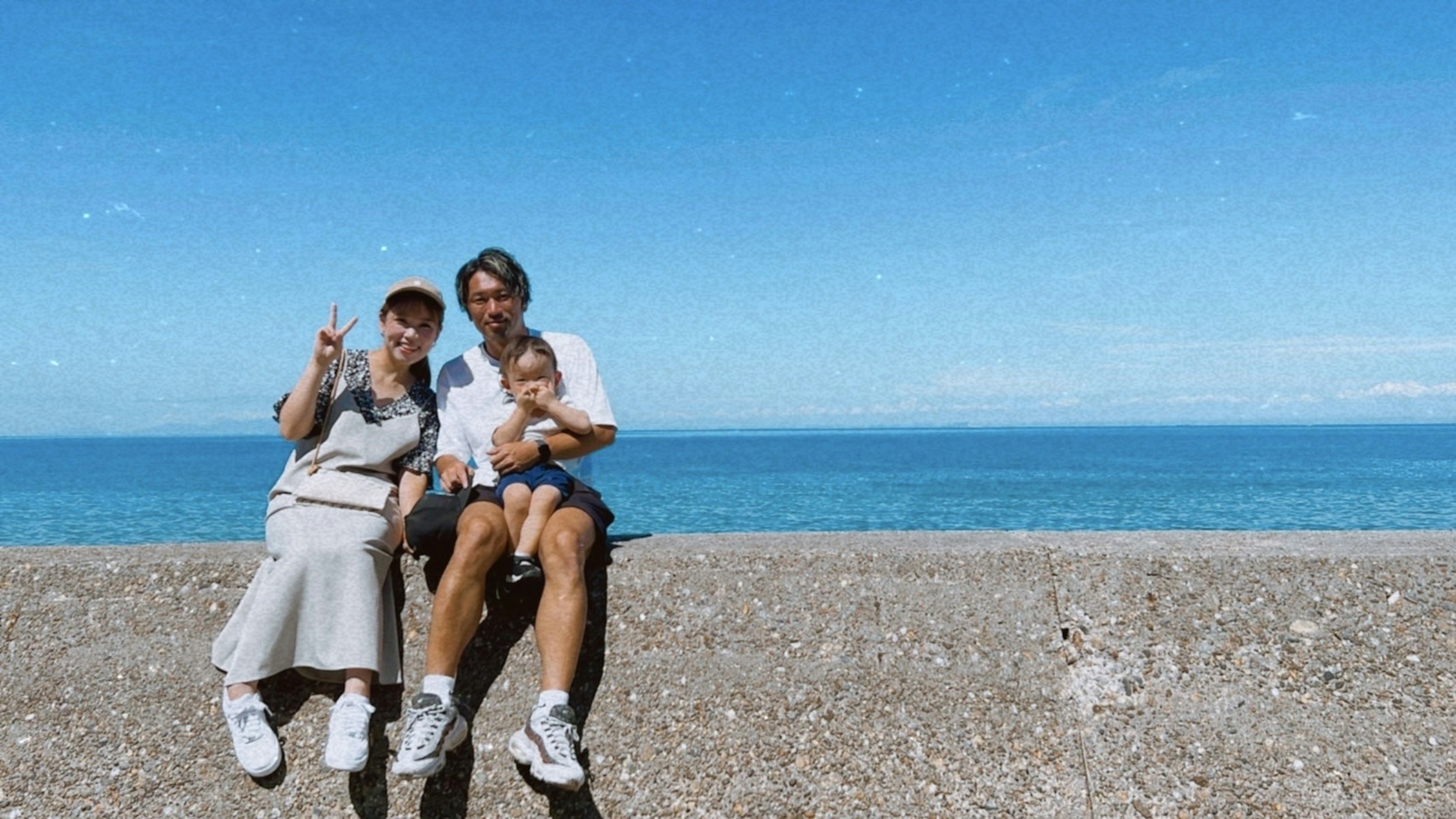 Foto keluarga dengan dua orang tersenyum dan bayi duduk di latar belakang laut biru dan langit