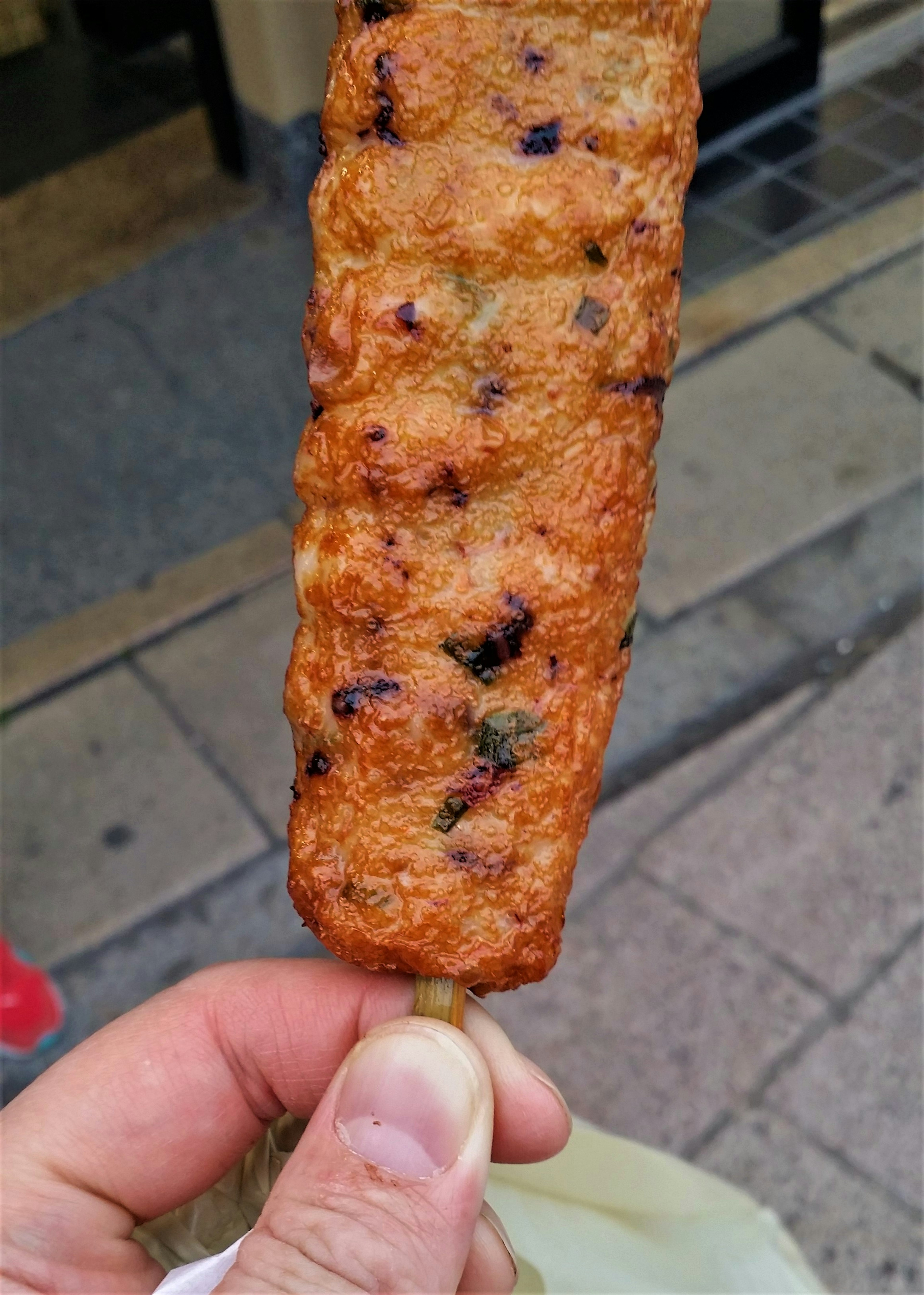 A hand holding a skewered fried snack with a crispy surface and dark specks