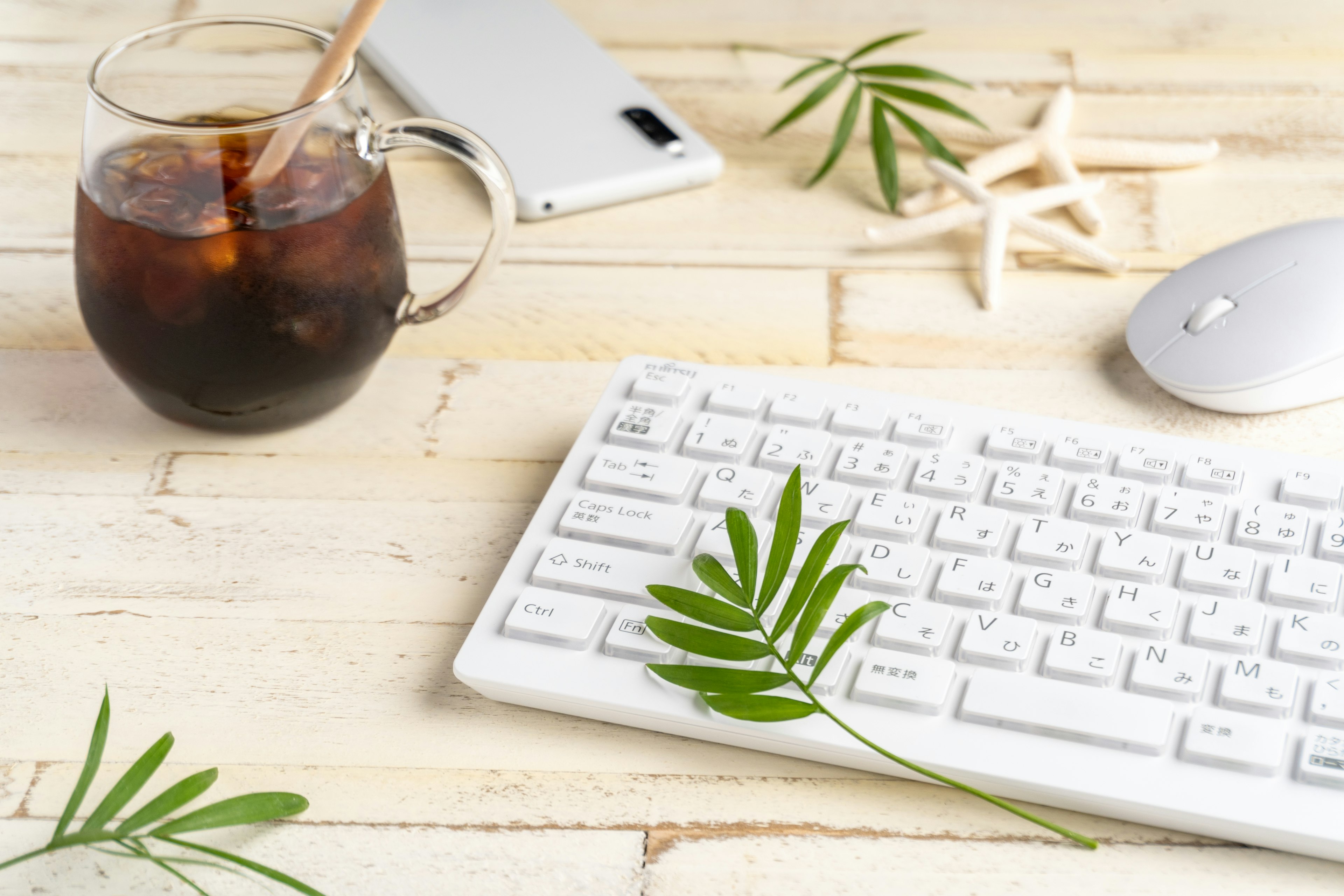 Scène de bureau avec un clavier et une souris sur une table en bois blanc, un verre de café glacé, des feuilles vertes et une étoile de mer