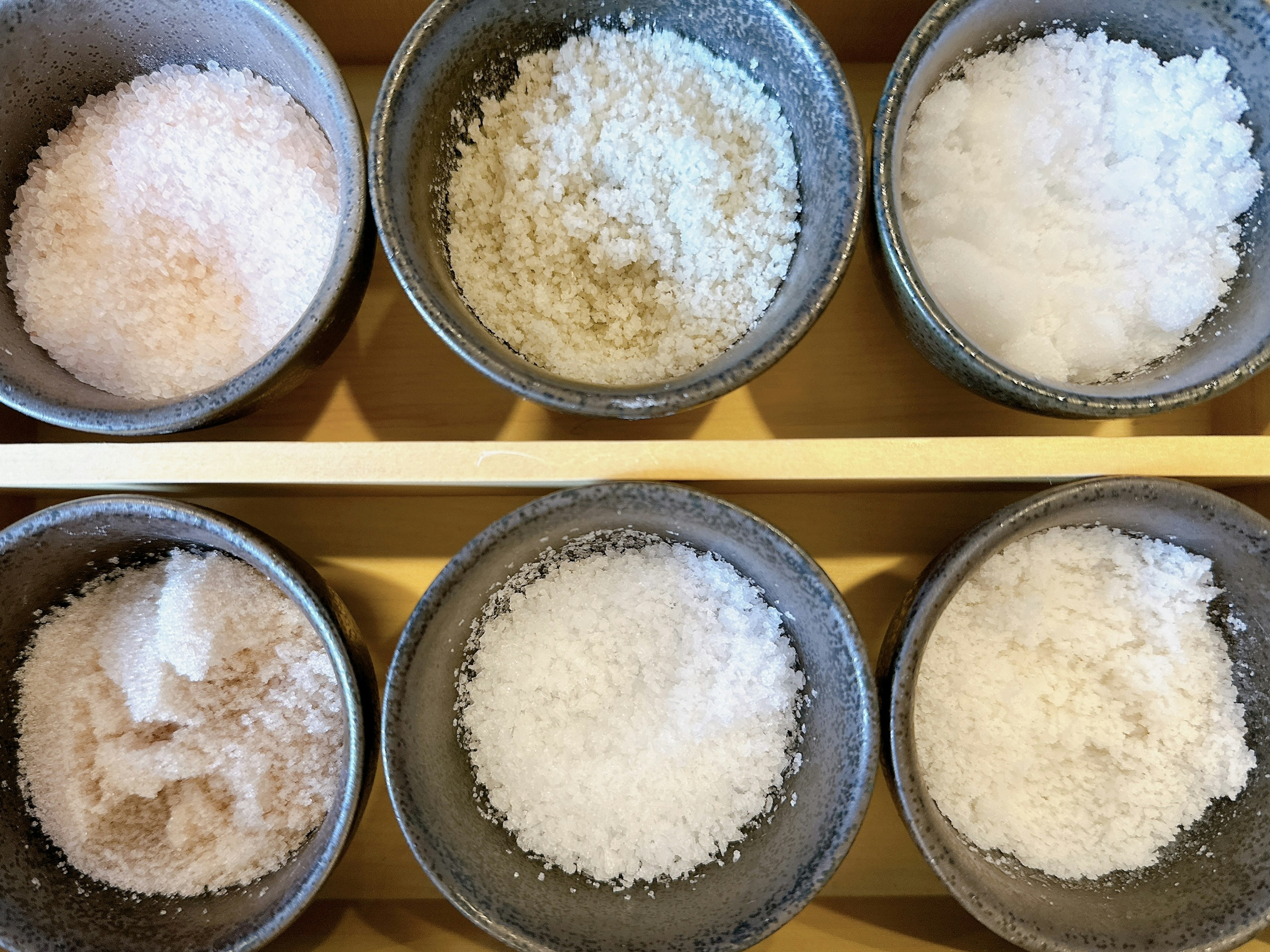 Six gray bowls filled with different types of salt arranged on a wooden tray