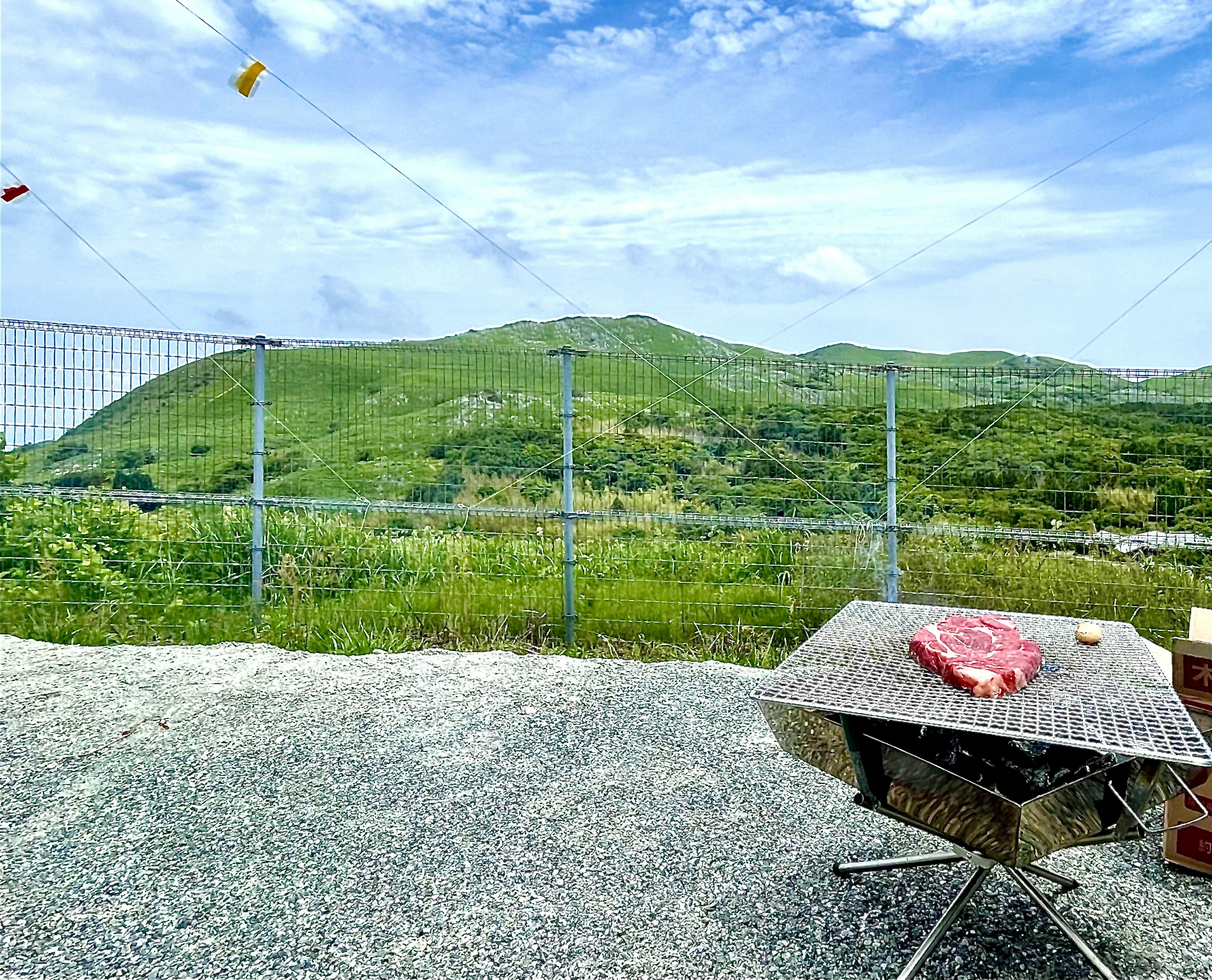 緑の丘と青い空が広がる風景に肉が置かれたバーベキュー台