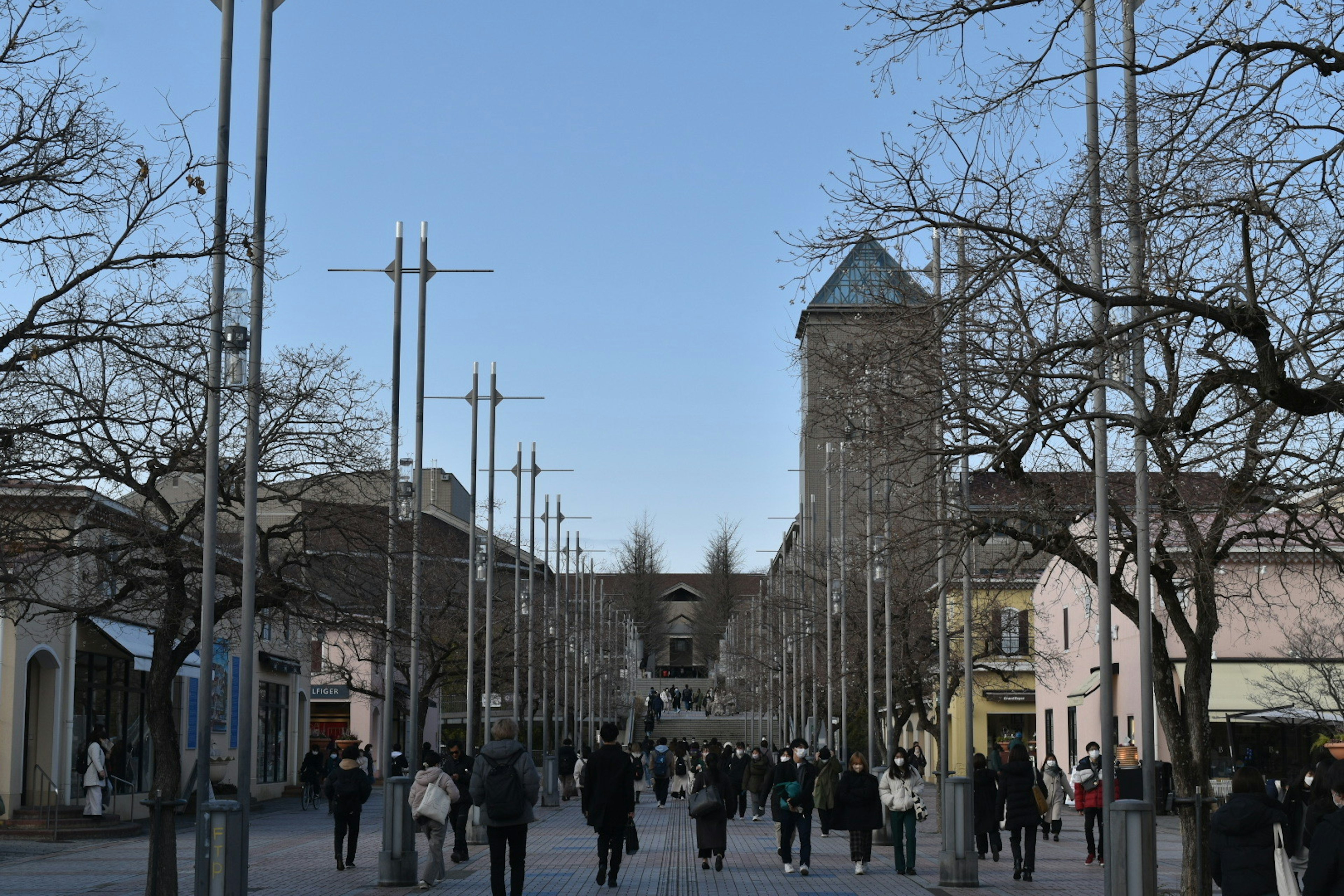 Personas caminando por una calle bajo un cielo azul con una torre al fondo