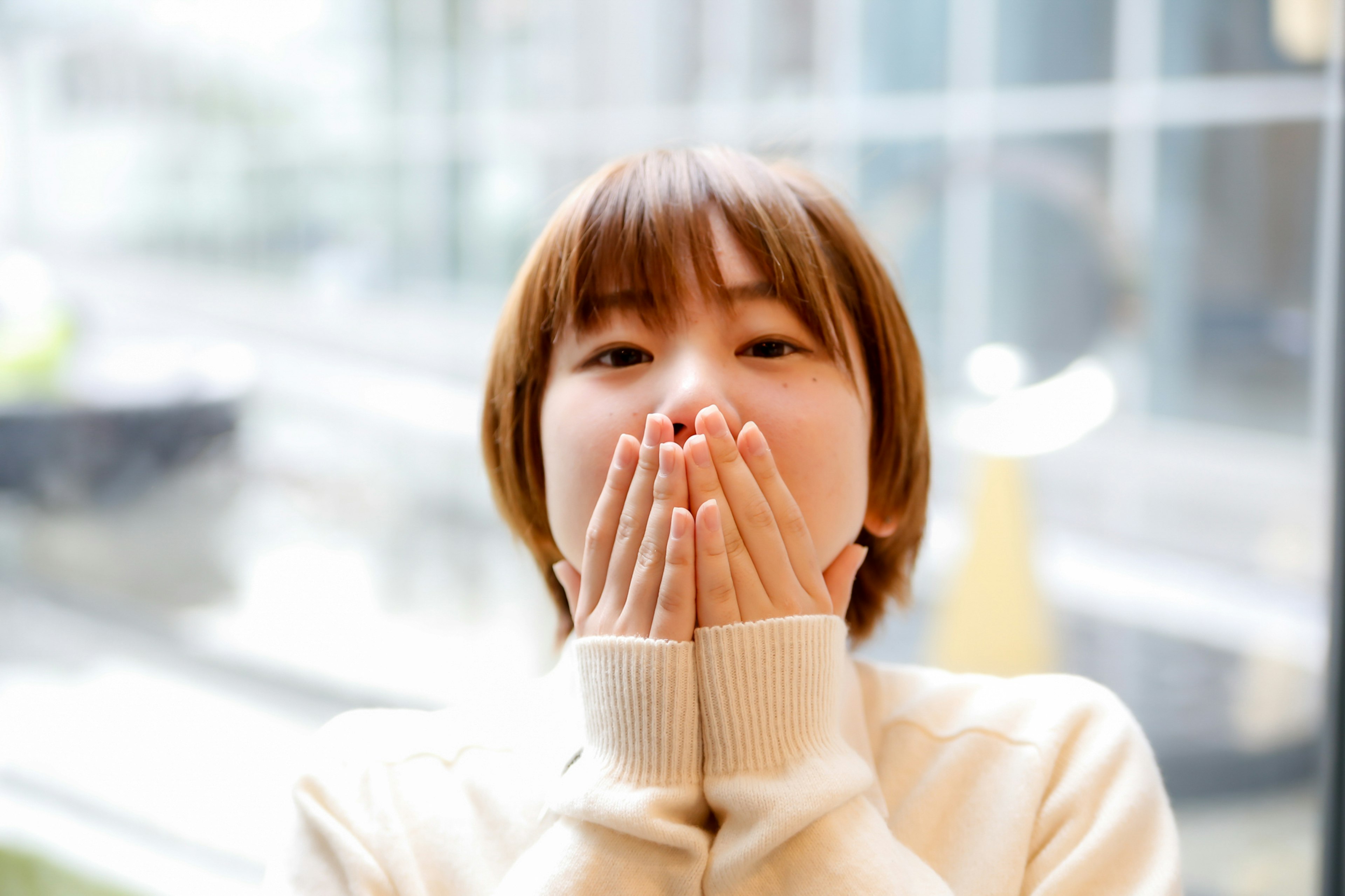 A woman expressing surprise or joy with her hands over her mouth wearing a sweater near a window