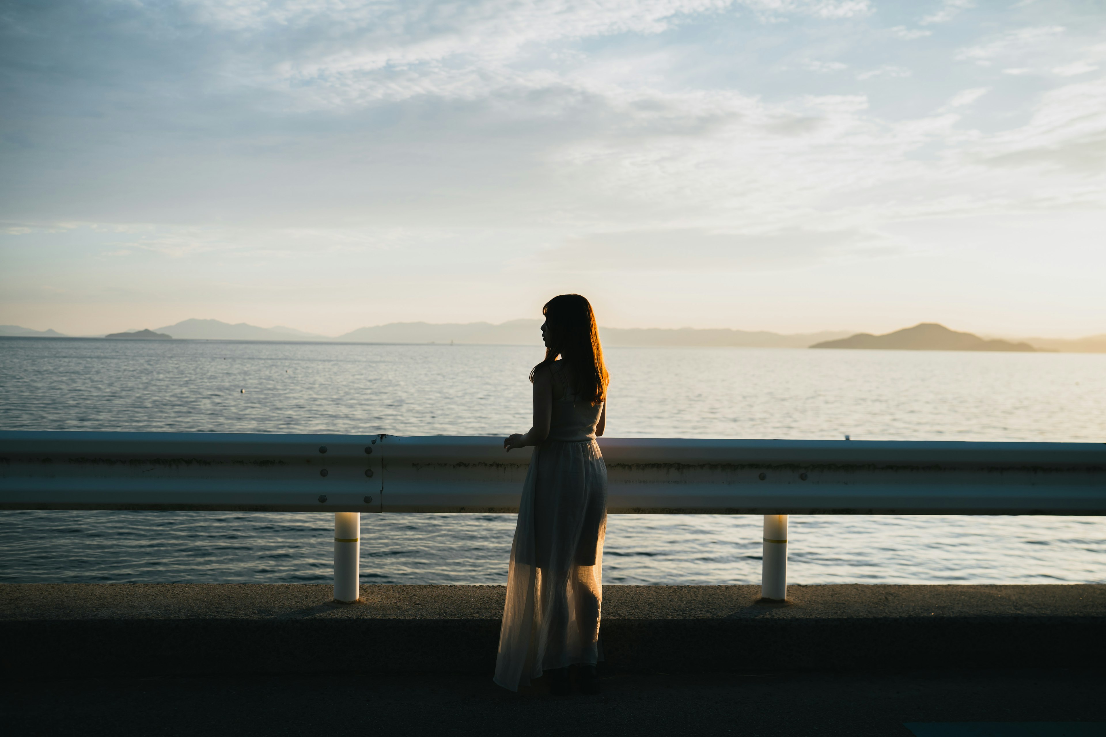 夕日を背景にした女性のシルエットが海を見つめる