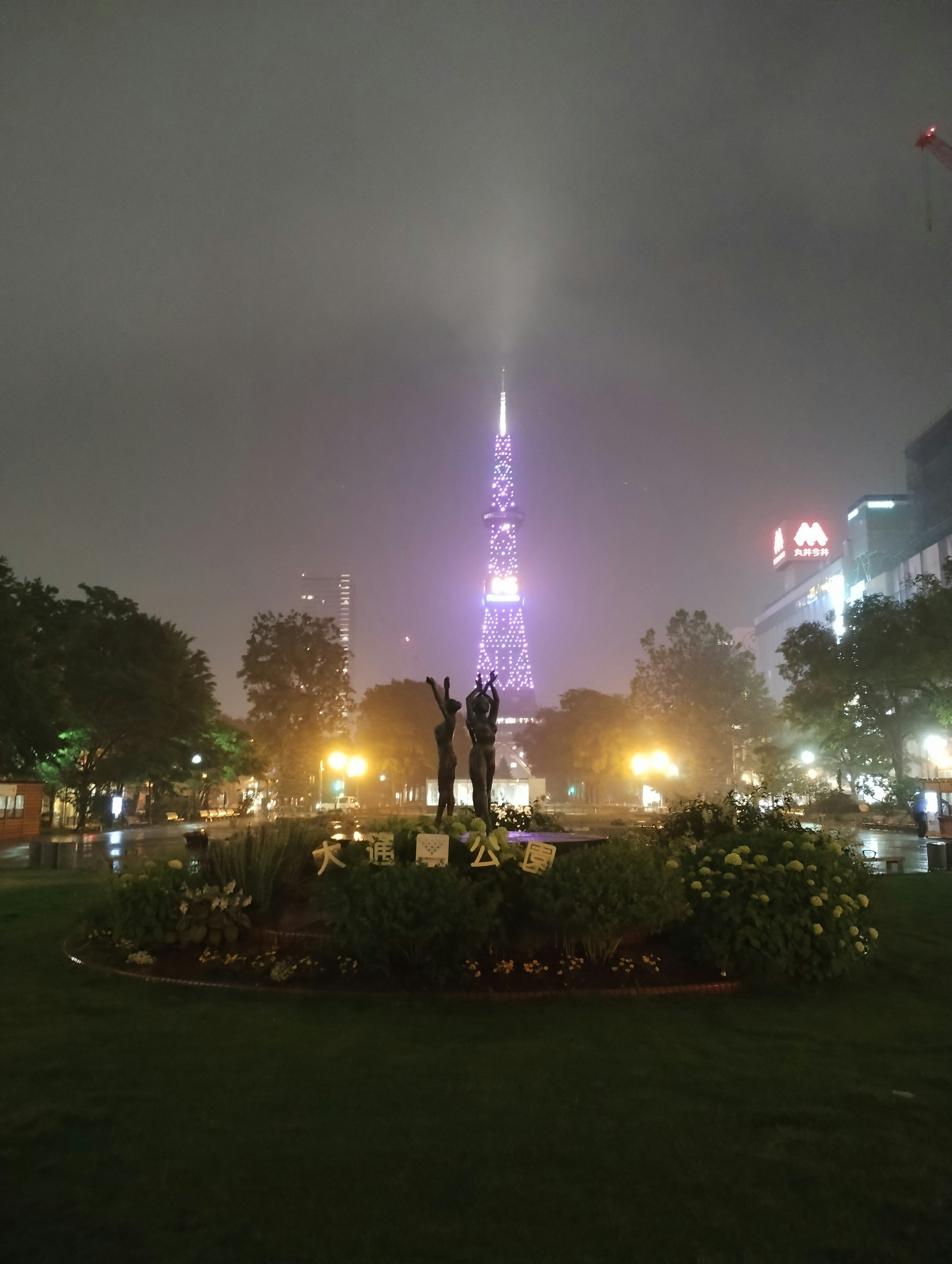 Park scene with a purple tower shrouded in fog and surrounding lights featuring a central statue