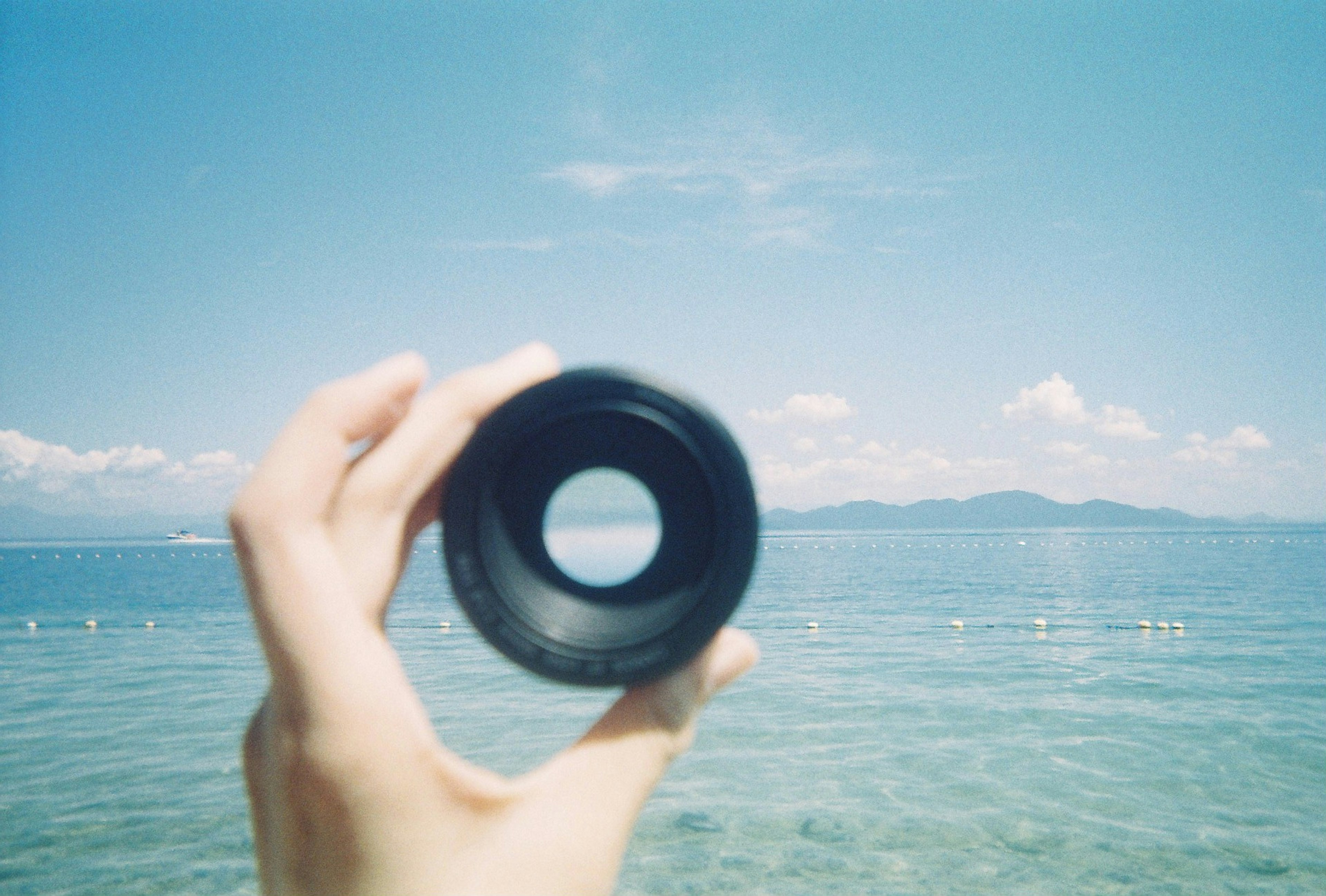 Une main tenant un objectif noir avec une vue sur la mer et le ciel bleus