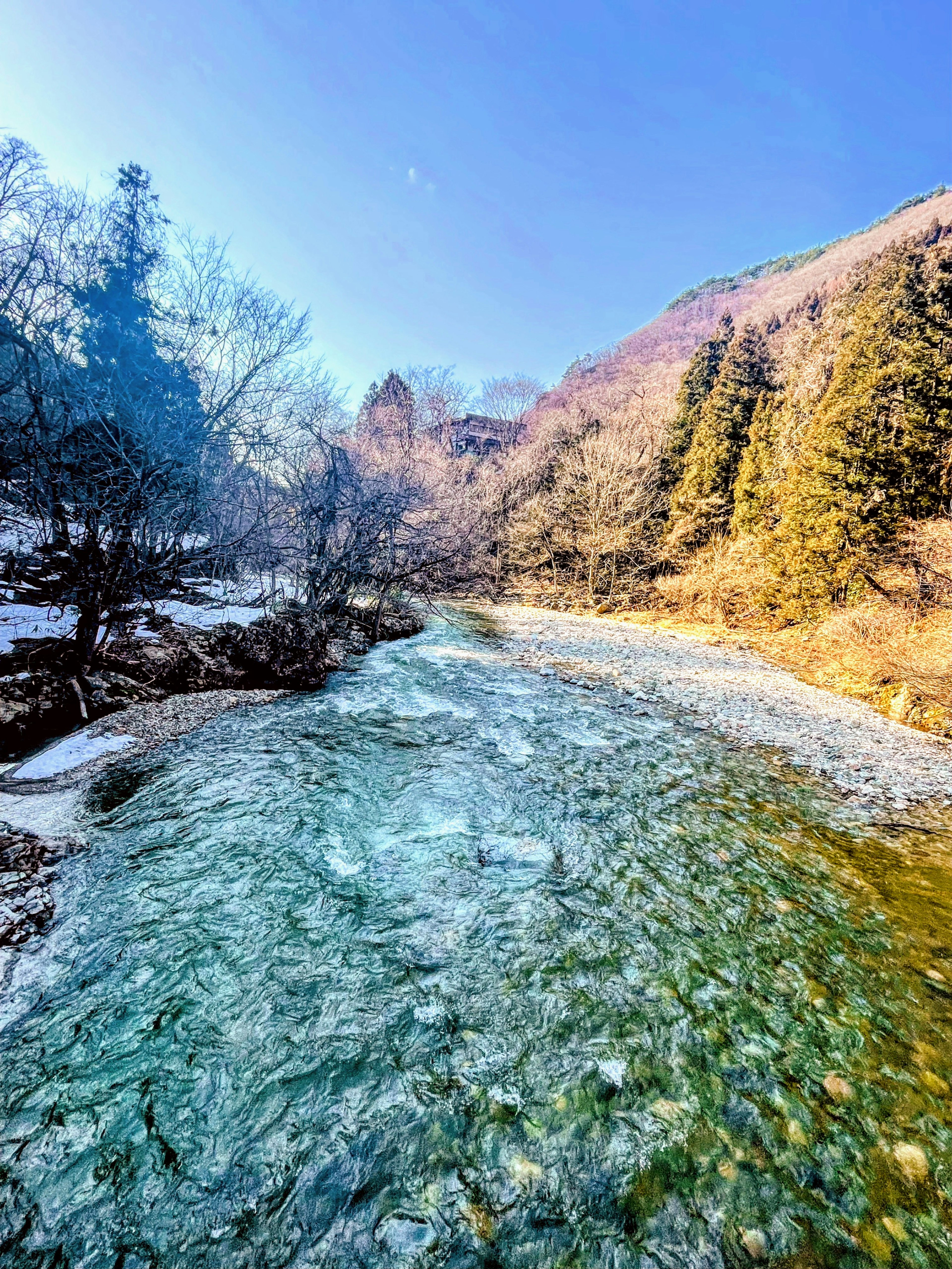 Vista panoramica di un fiume con residui di neve circondato da montagne e alberi sotto un cielo blu