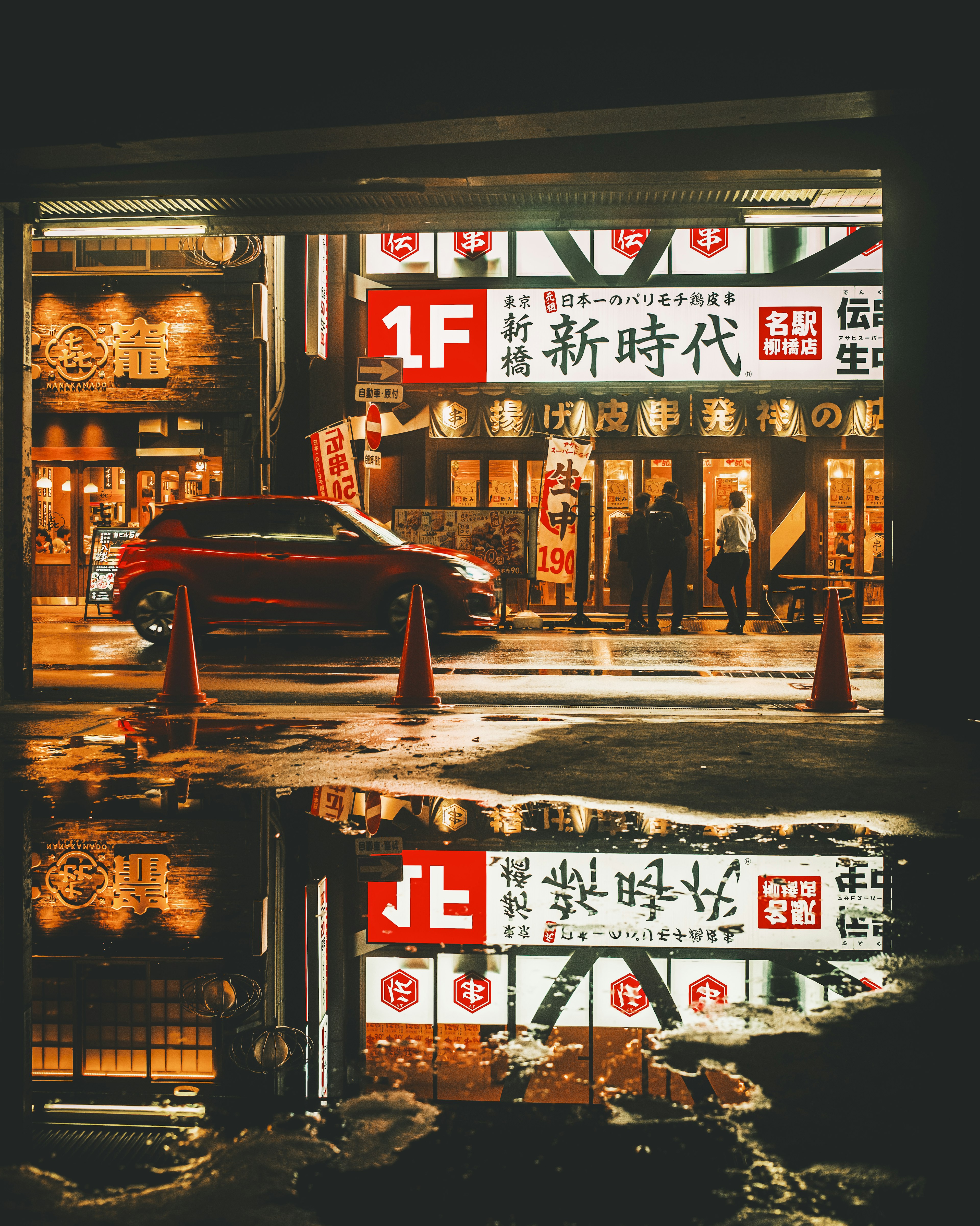 Night city scene with a red car and neon signs reflected in water