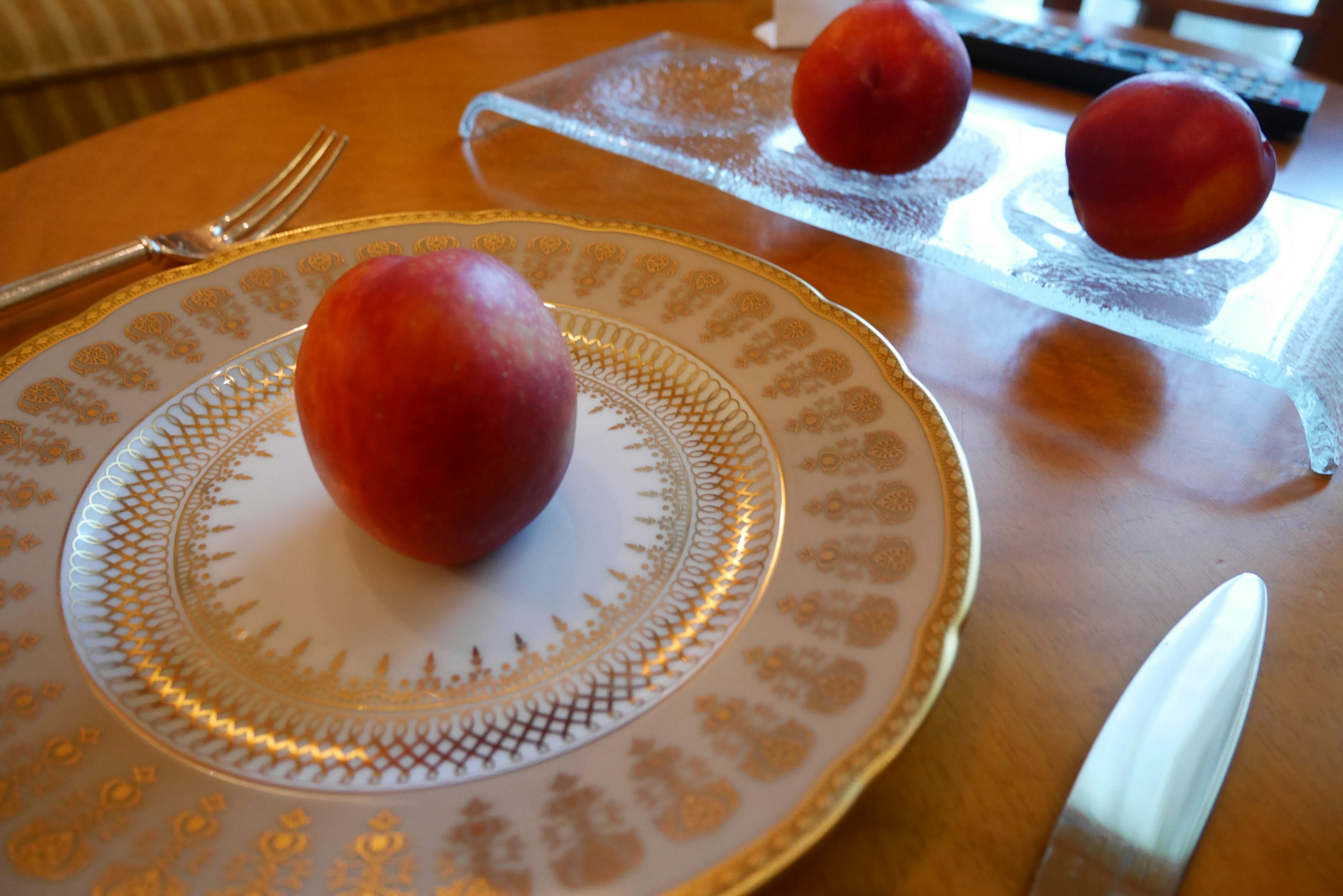 Un durazno rojo sobre un plato decorado con borde dorado en una mesa de madera con más duraznos en una bandeja de vidrio