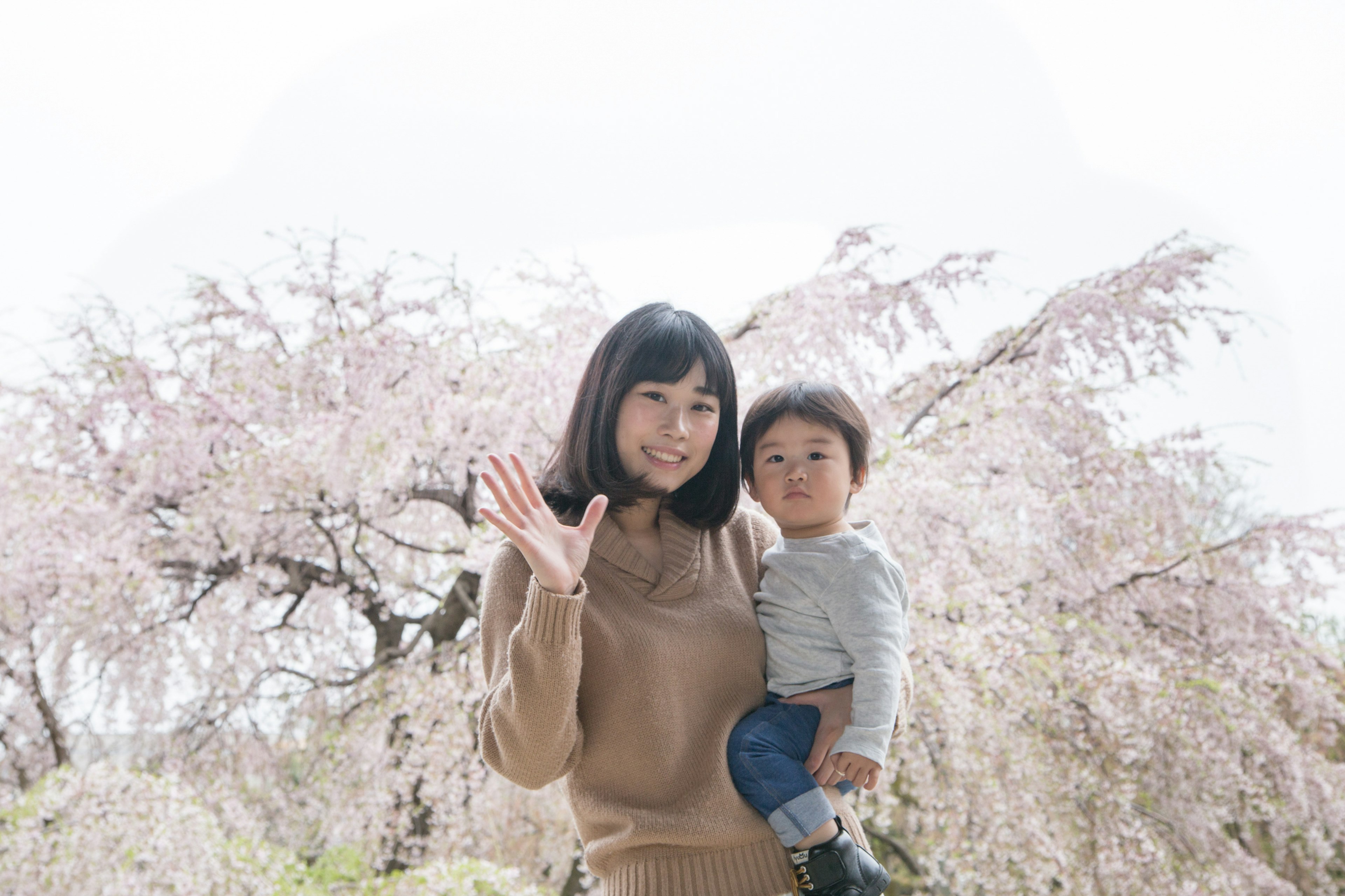 Une femme faisant un signe de la main avec un enfant dans ses bras devant des cerisiers en fleurs