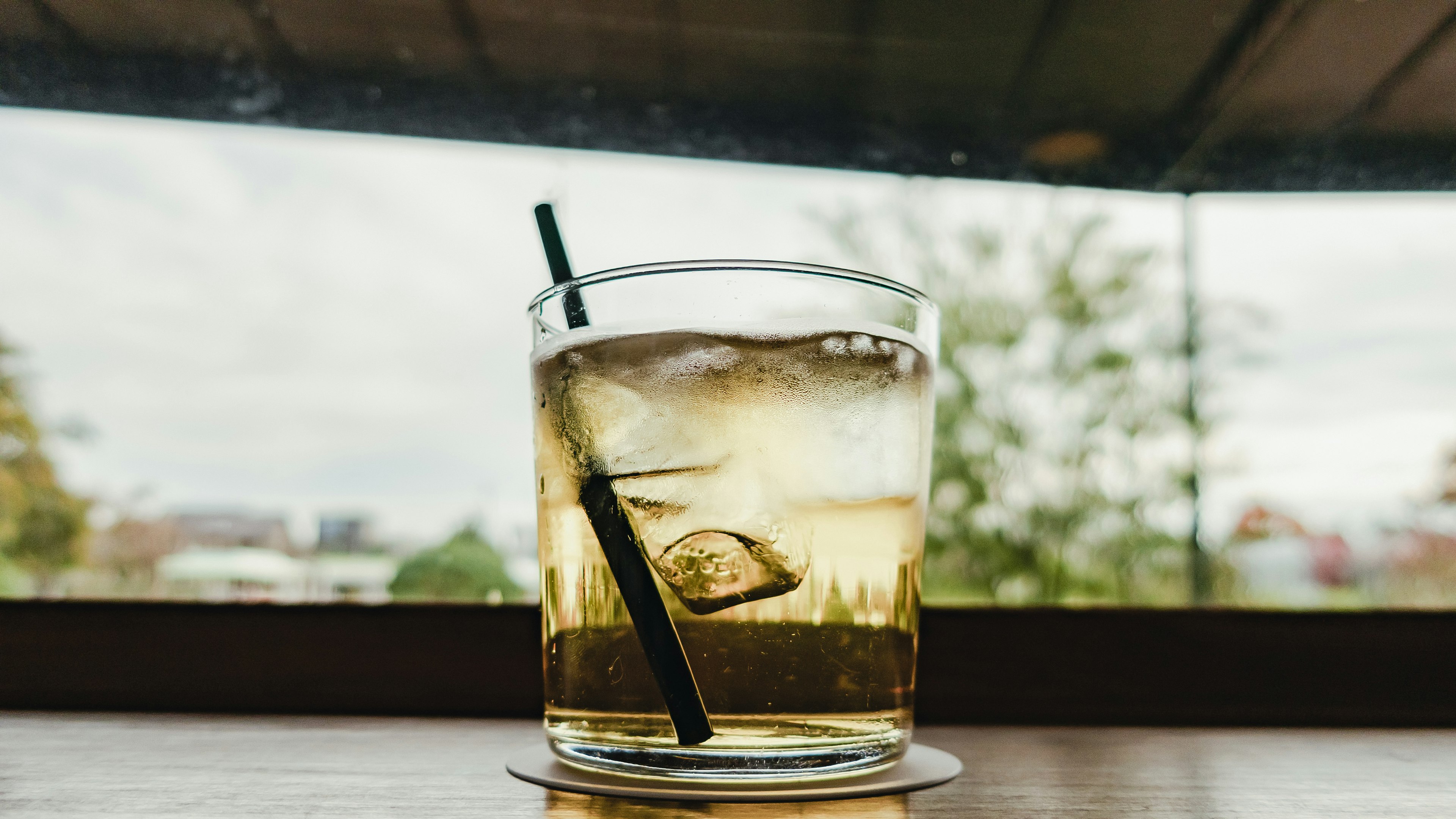 Whiskey glass with ice and a black straw against a backdrop of green trees visible through a window