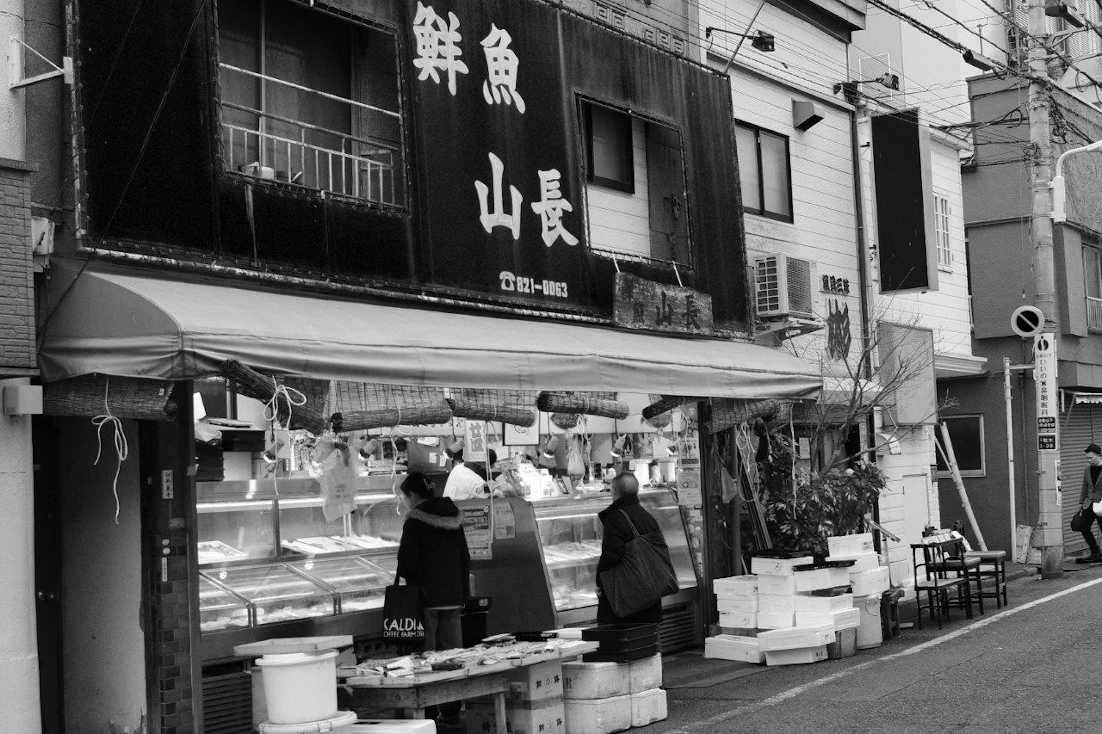 Image en noir et blanc de l'extérieur d'un marché aux poissons avec un panneau indiquant 'Poisson frais Yamacho'