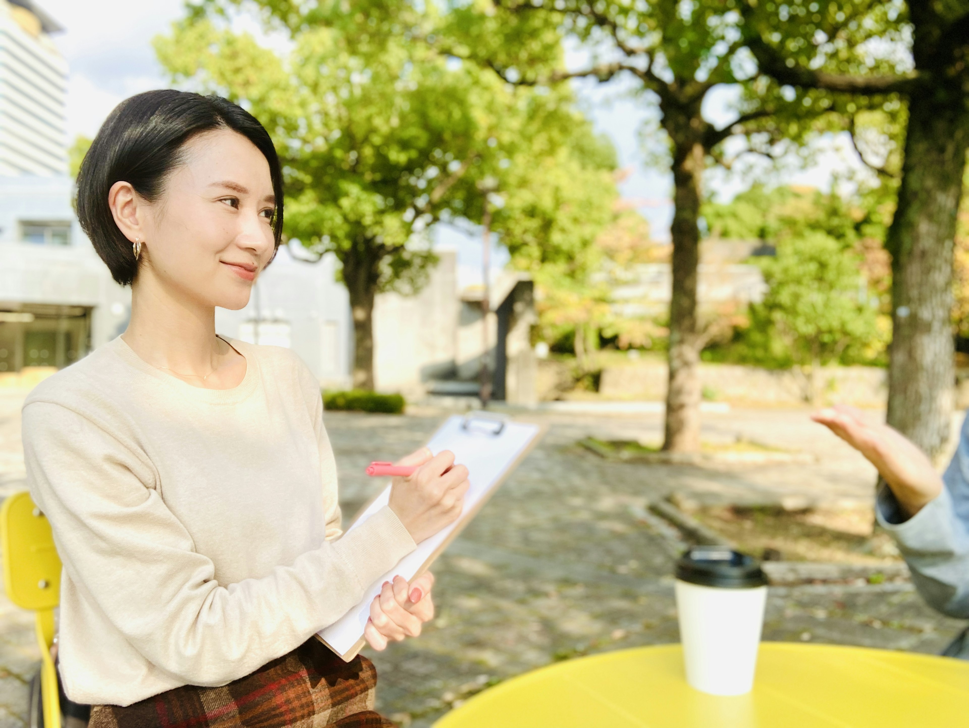 Woman being interviewed in a park holding a clipboard at a yellow table with green trees in the background