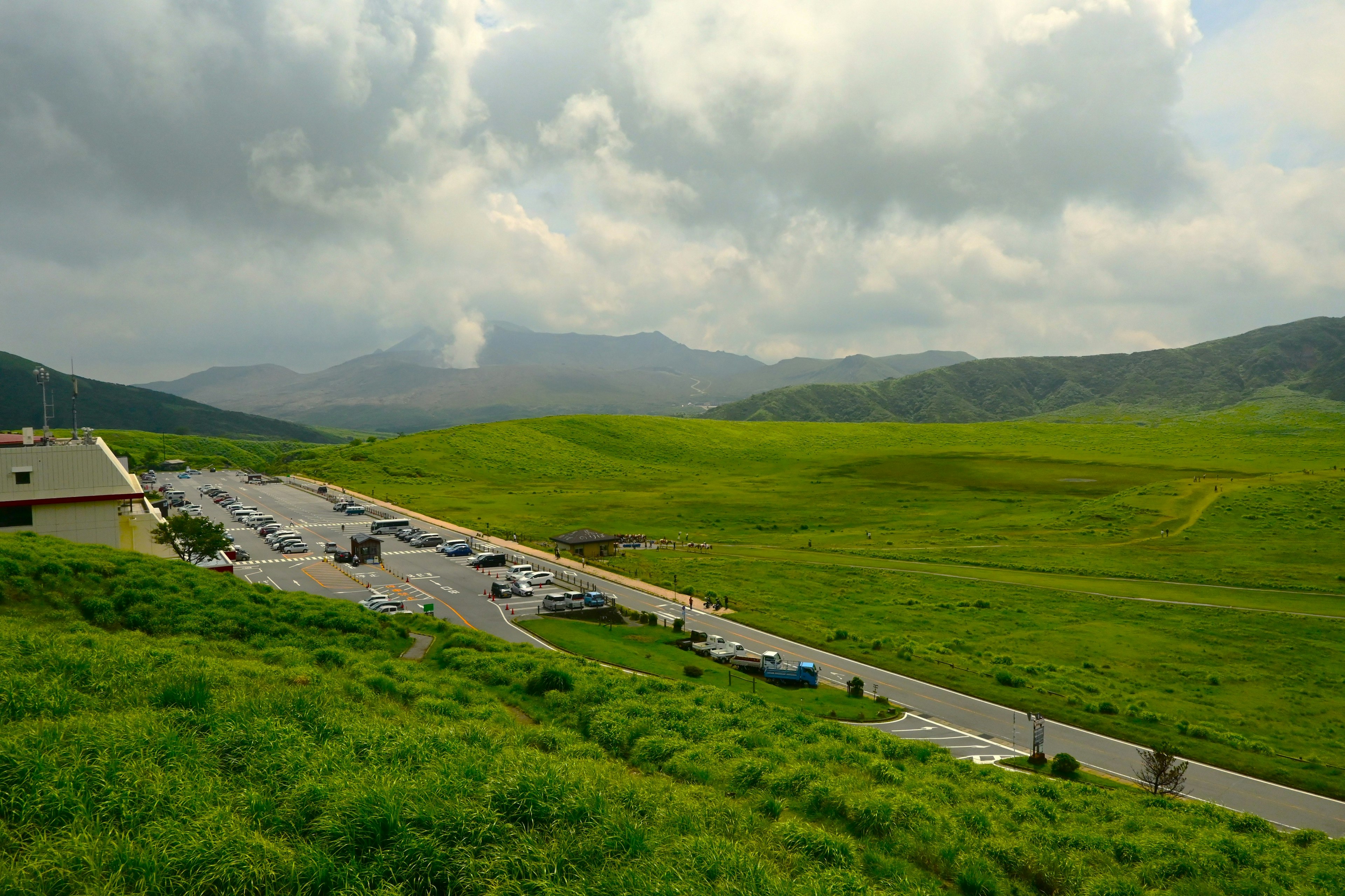 美しい緑の草原と駐車場が広がる風景、雲が多い空と遠くの山々